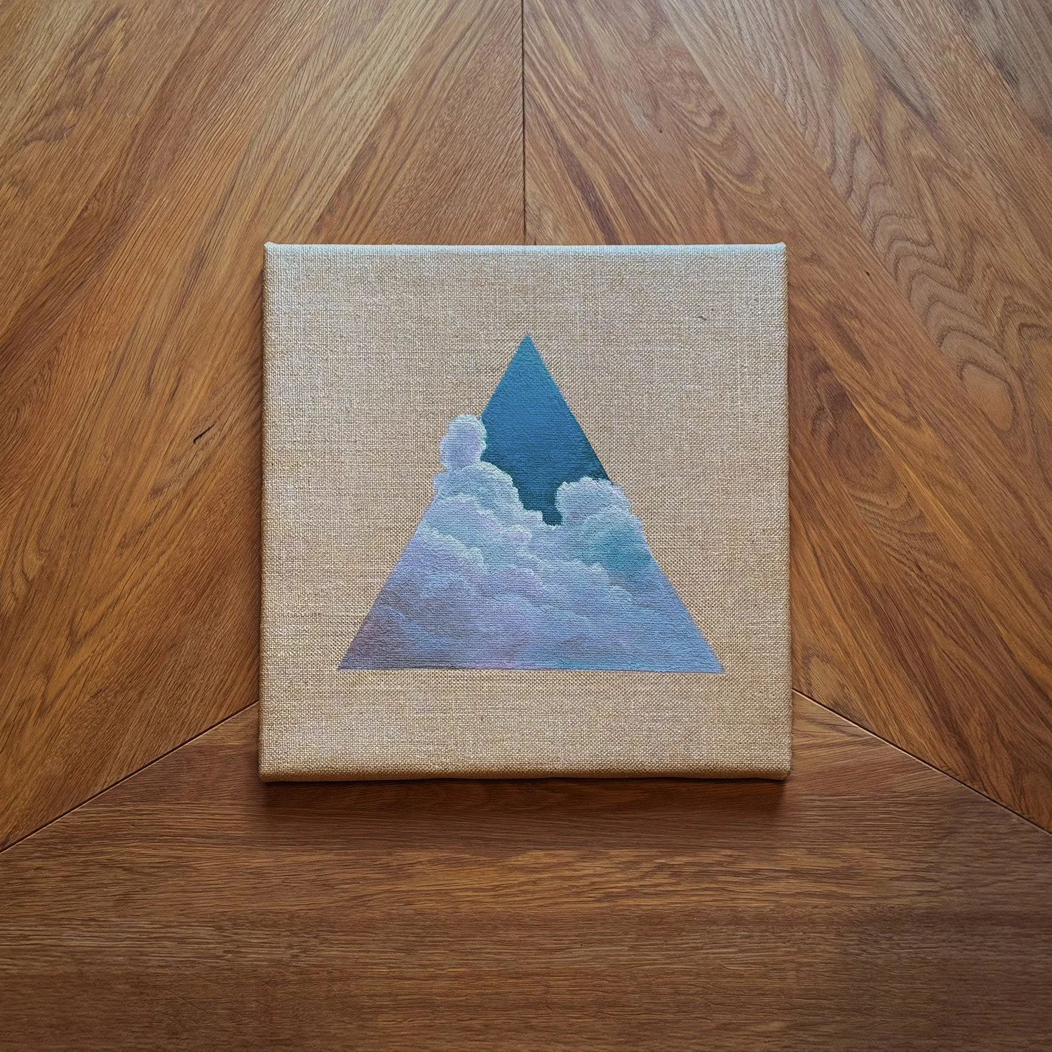 Ein Gemälde zeigt eine blaue Pyramide mit Wolken im inneren und im Himmel. Es liegt auf einem Holztisch.