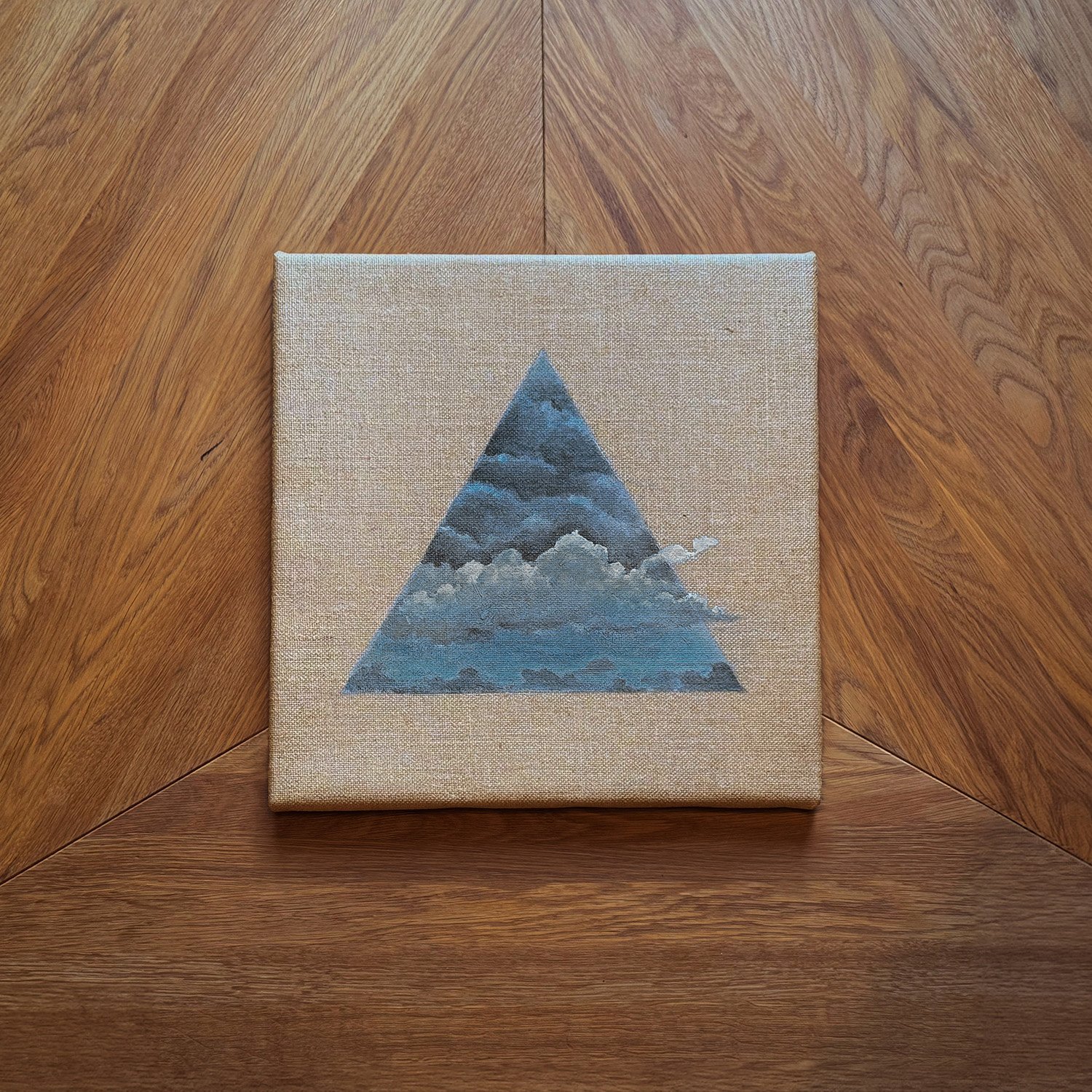 Ein quadratisches Kunstwerk mit einer Dreiecksform, die Wolken im Himmel zeigt, auf einem Holztisch.