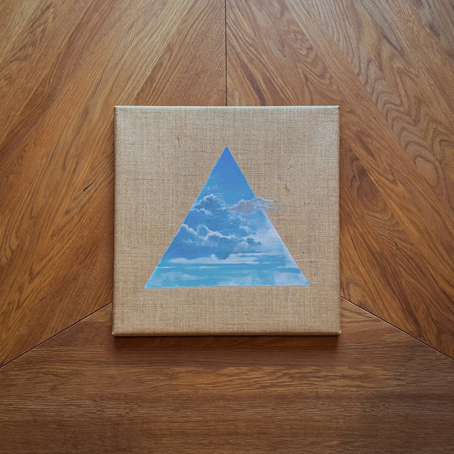 Ein Kunstwerk mit einem blauen Dreieck, das einen Himmel mit Wolken zeigt, auf einem Leinwand auf einem Holztisch.