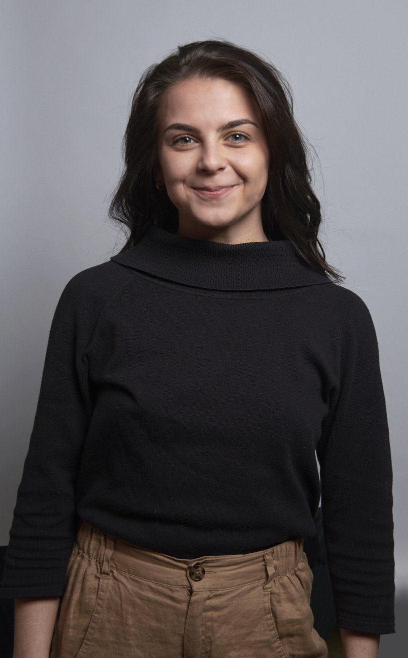 A young woman with shoulder-length brown hair, smiling and wearing a black long-sleeve top and tan pants against a light gray background.