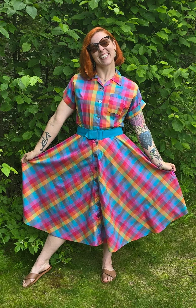 A girl standing outside wearing sunglasses, smiling, and wearing a vintage rainbow plaid dress