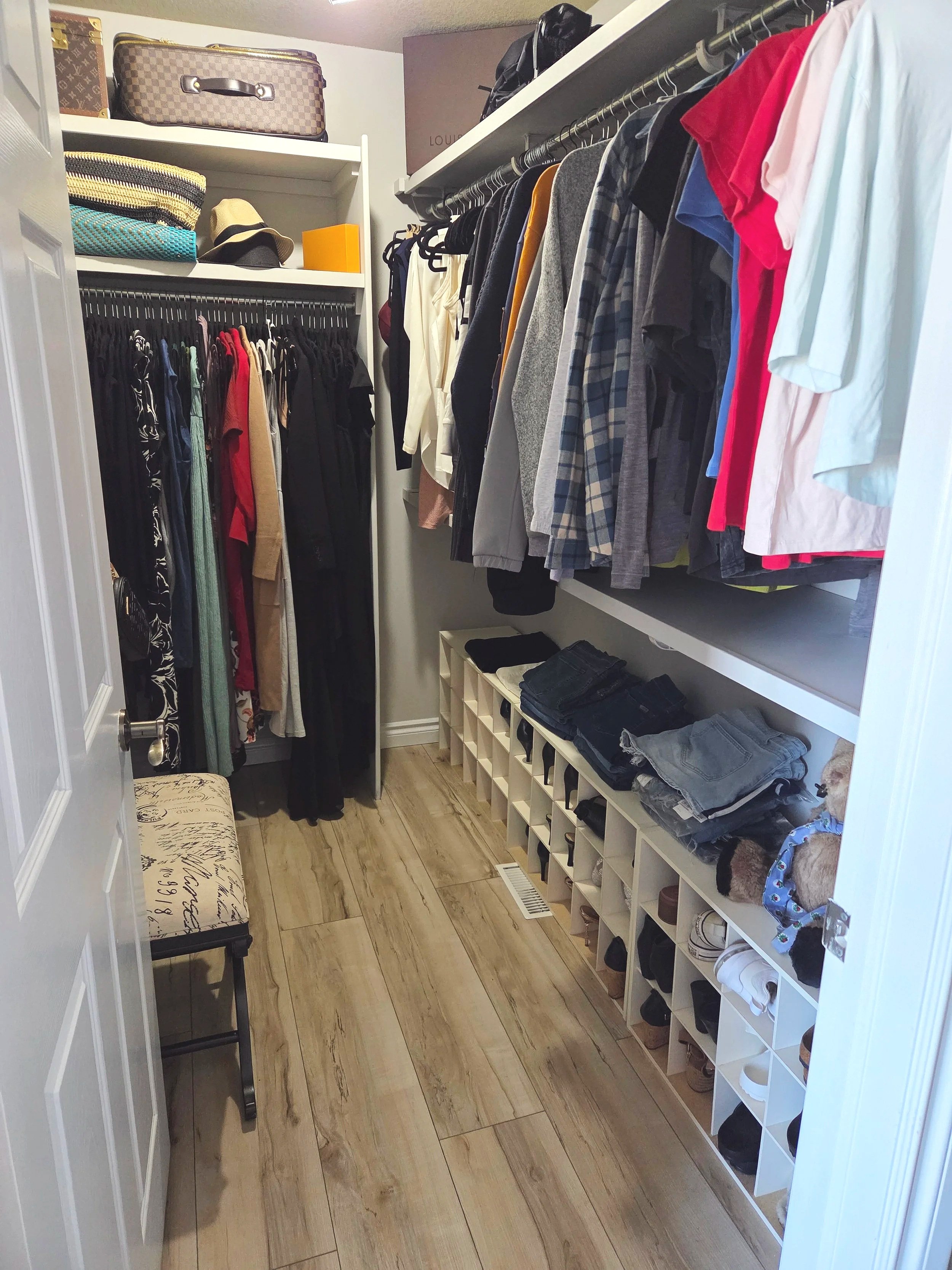 A walk-in closet with shelves and hanging rods filled with clothes, shoes, handbags, and other accessories. A small bench with a cushion is on the left, and there are suitcases and boxes on the top shelves.