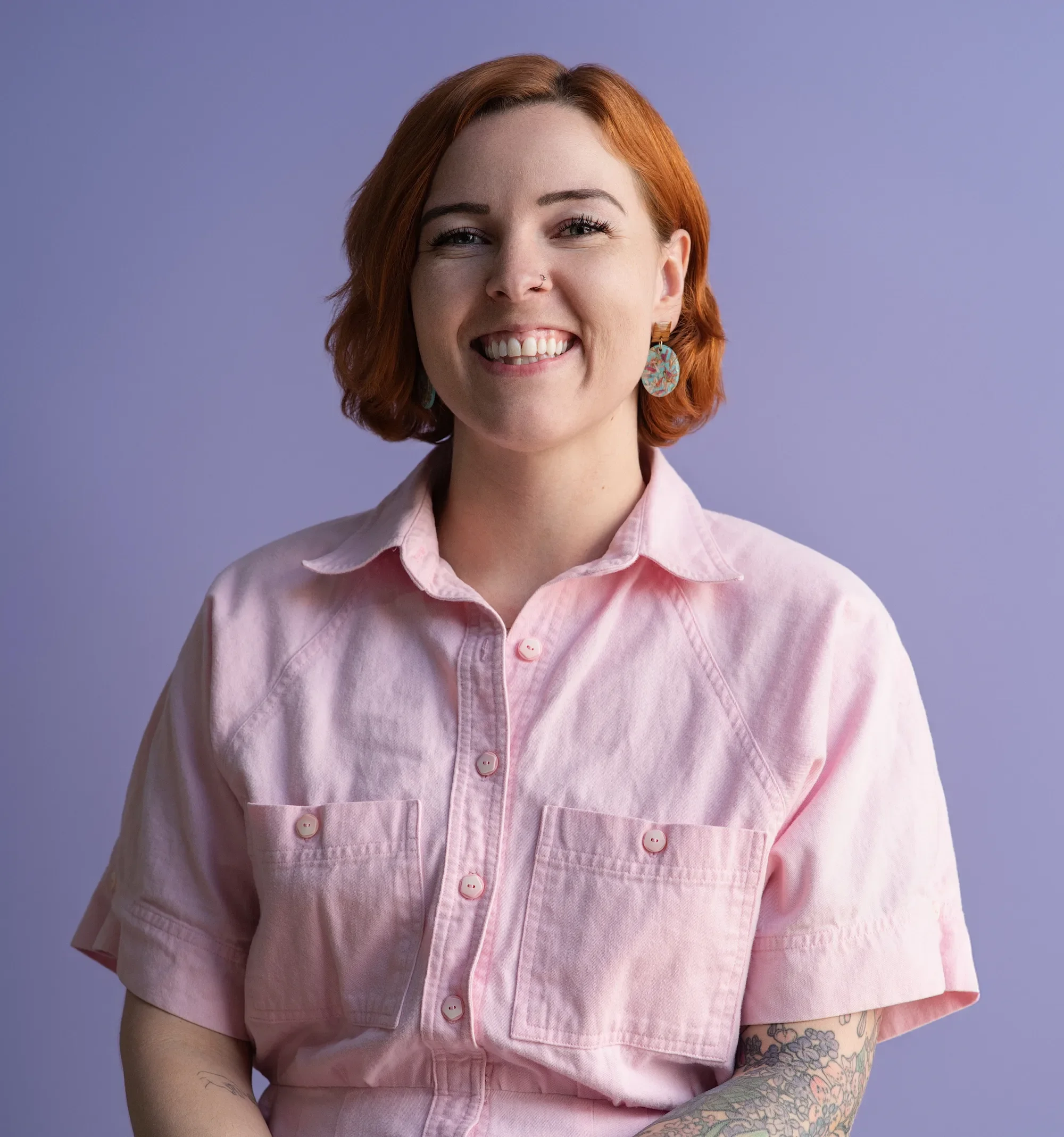 Image of a smiling, red headed woman wearing a pink vintage dress against a purple backdrop