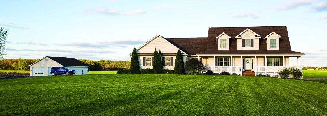Large white house with a wraparound porch, multiple dormer windows, surrounded by a green lawn, with a detached garage and a blue car parked outside.