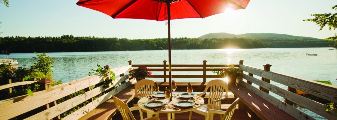 A lakeside outdoor dining setup with a round table, four chairs, and a large red umbrella on a wooden deck, overlooking a lake with trees and mountains in the background.