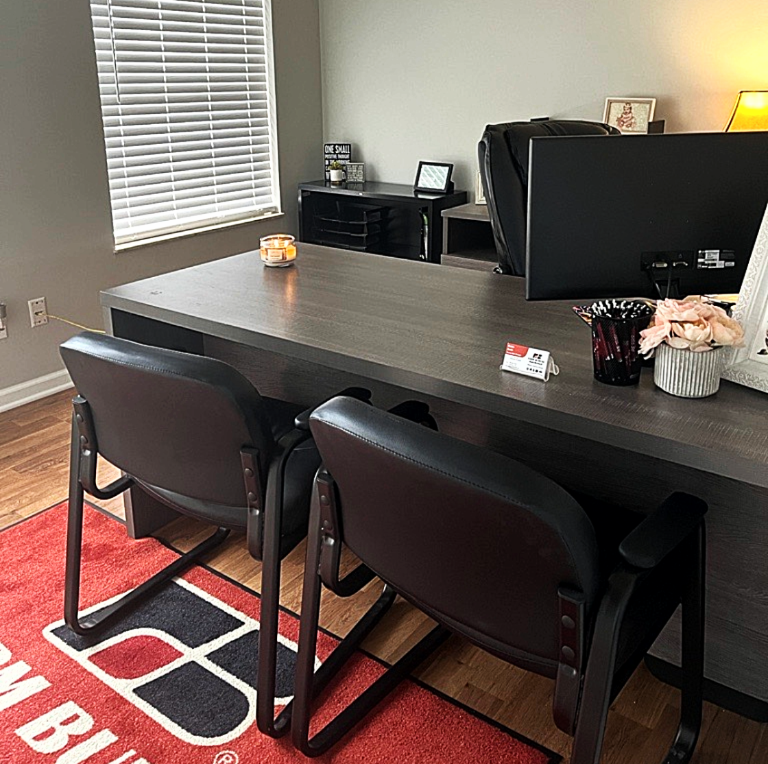 Office space with a large desk, two black chairs, a computer monitor, a vase with flowers, a candle, and a window with blinds.