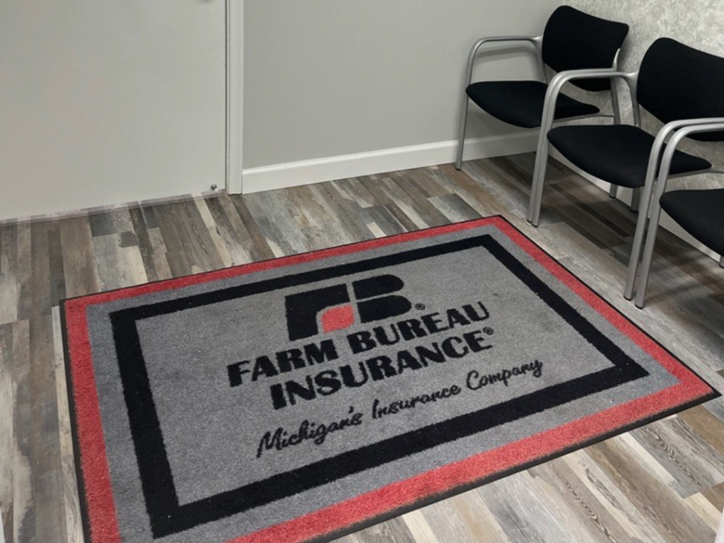 Indoor waiting room with three black chairs, a gray wall, a door, and a gray, red, and black FARM BUREAU INSURANCE mat on wood-look flooring.