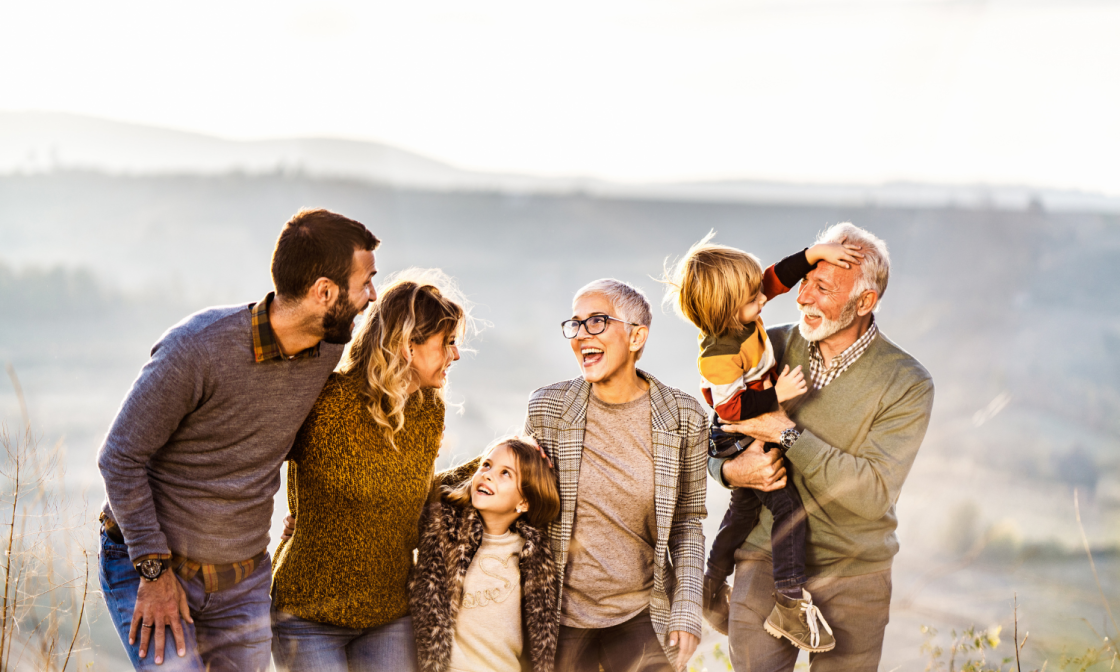 A multigenerational family outdoors enjoying sunny weather, smiling and interacting with each other, with a scenic landscape in the background.