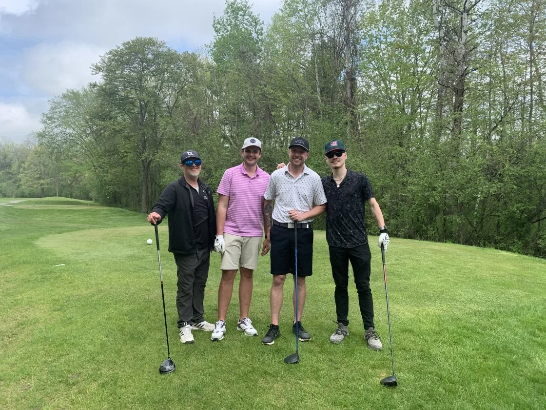 Four men standing on a golf course holding golf clubs, smiling at the camera, with trees and a cloudy sky in the background.