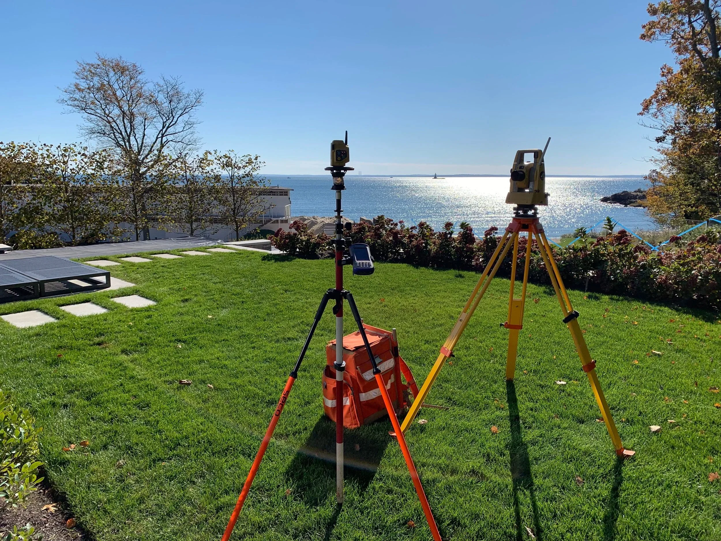 Surveying equipment set up on a grassy area overlooking a body of water with trees, a building, and a distant boat in the background during daytime.