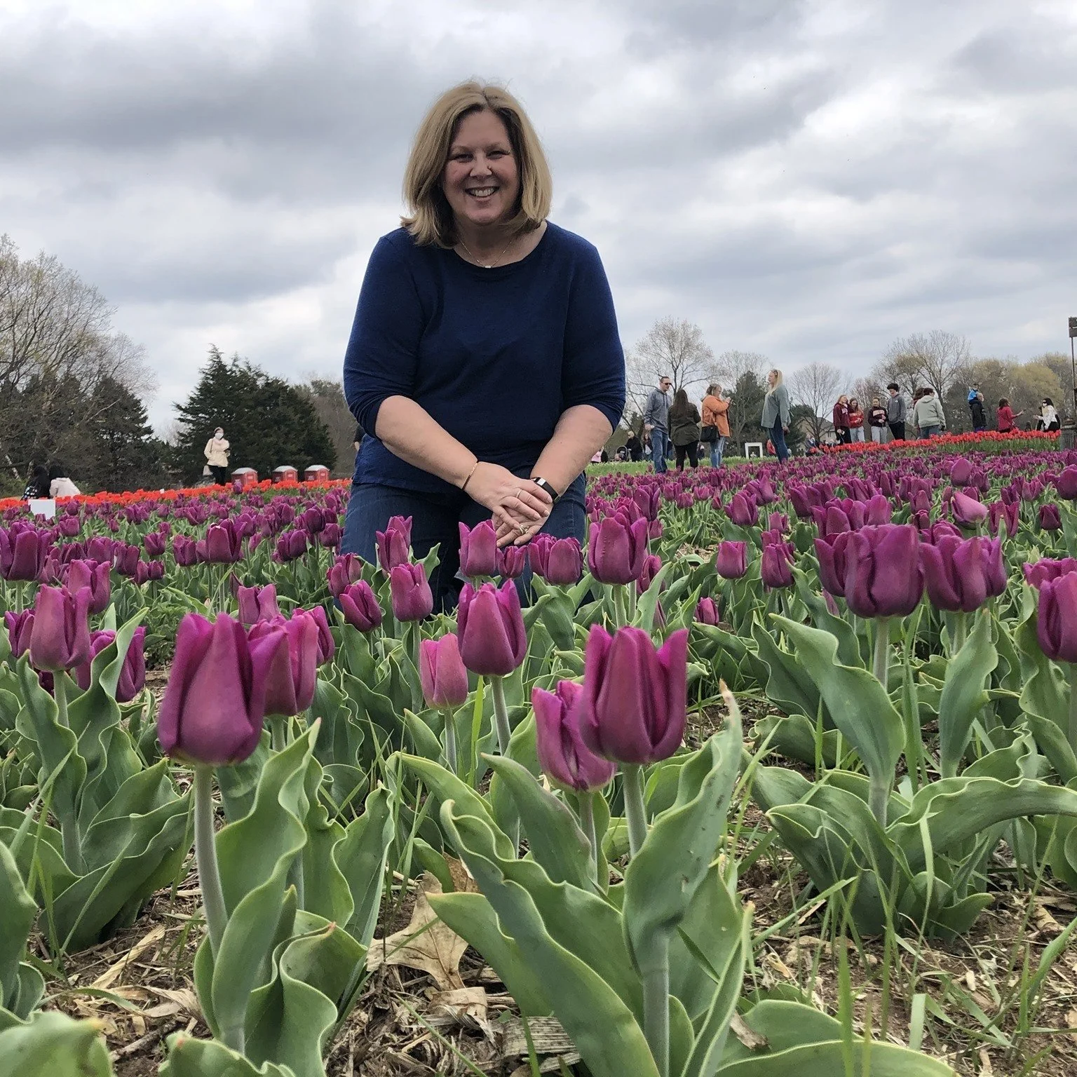 A smiling woman with shoulder-length blond hair kneels among purple tulips in a flower field, with people walking in the background on a cloudy day.