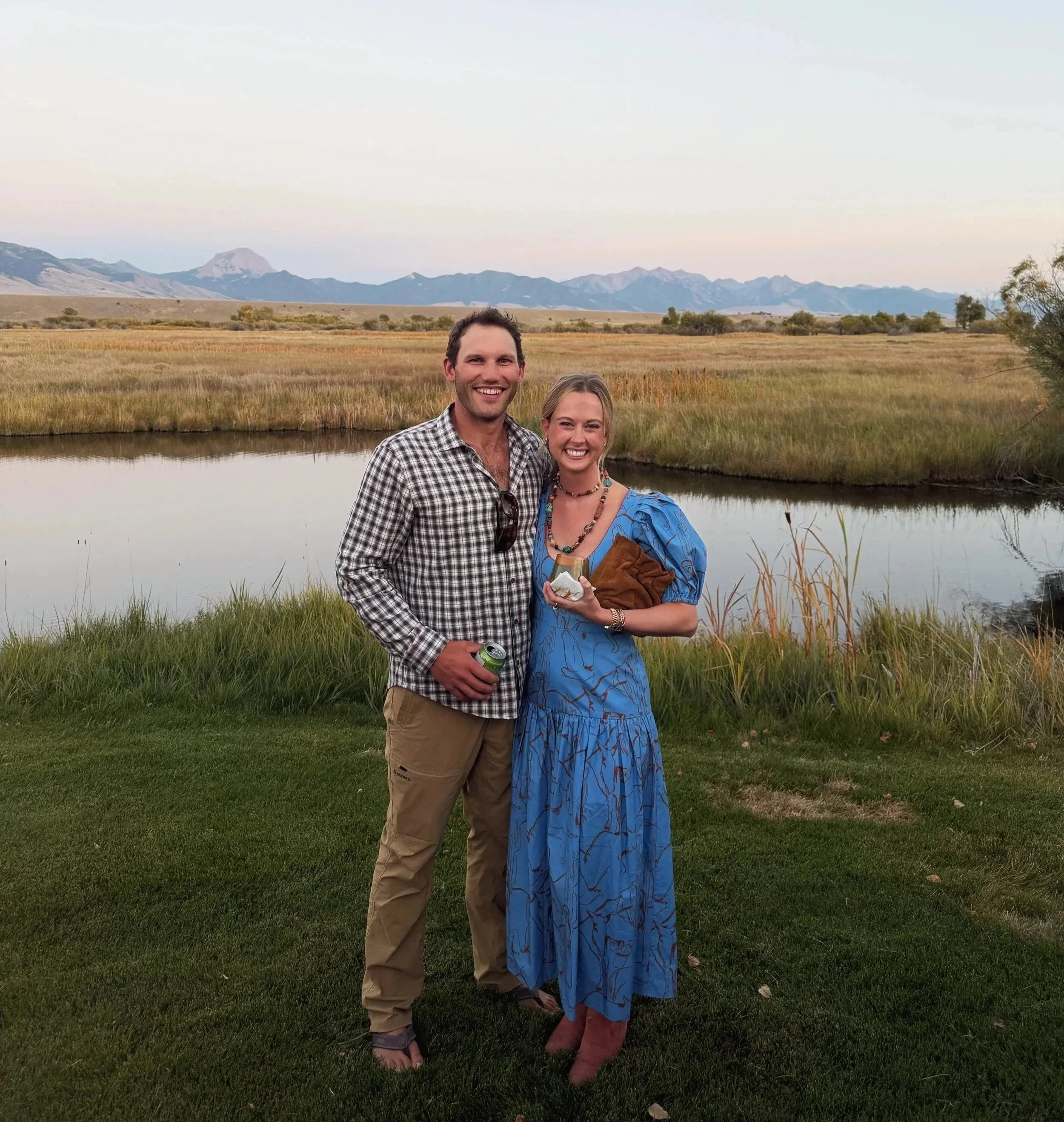 A smiling man and woman standing outdoors near a river with mountains in the background during sunset. The man is wearing a checkered shirt and khaki pants, holding a soda can. The woman is wearing a blue dress with a leaf pattern, holding some jewelry and a brown purse.