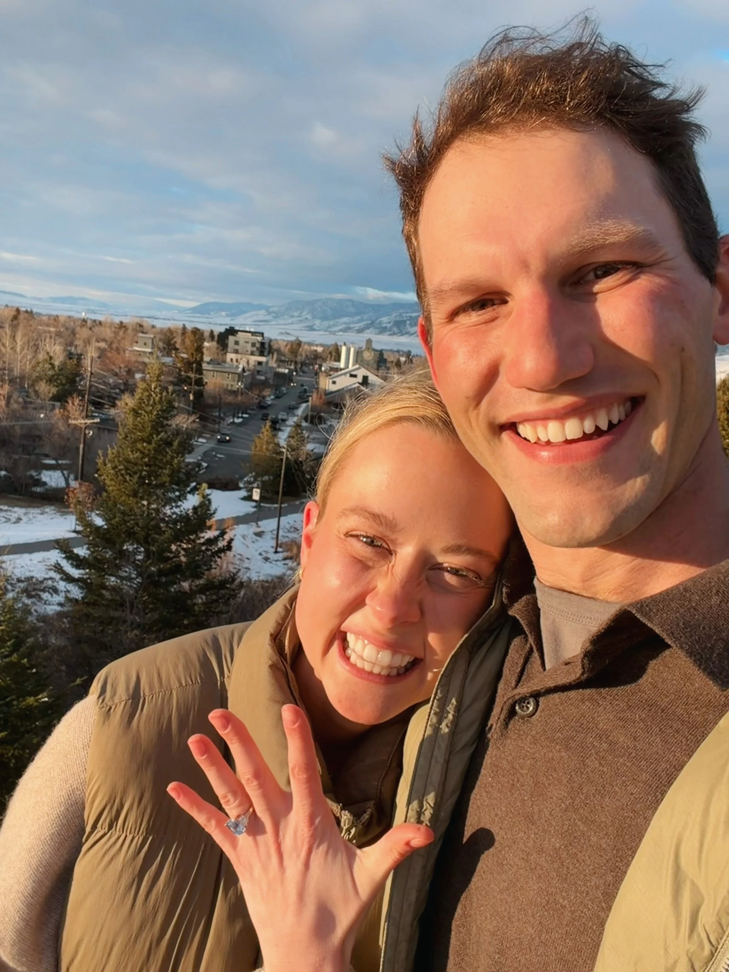 A smiling man and woman taking a selfie outdoors with a snowy landscape and mountains in the background.