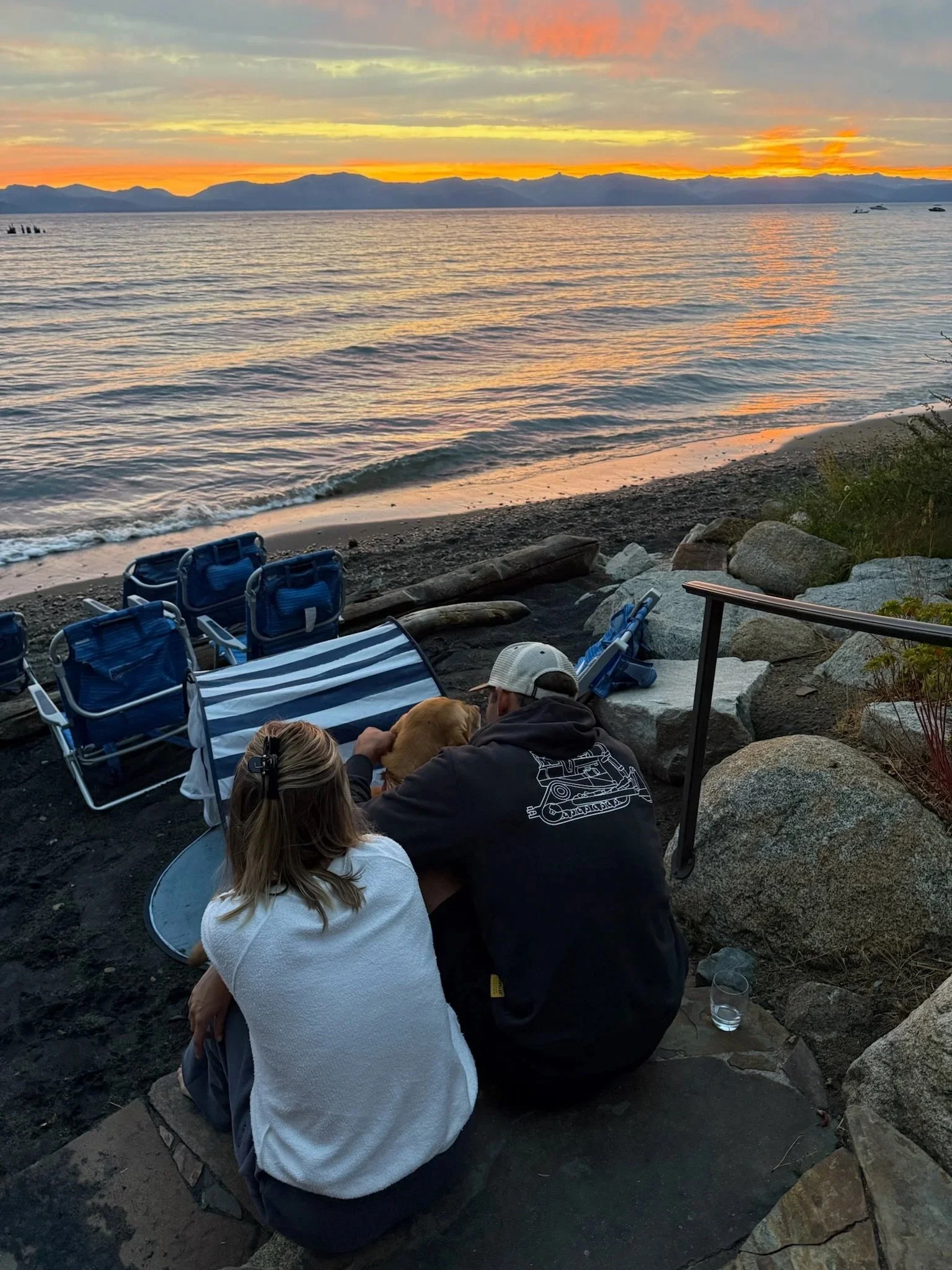 A couple sitting on a rock by the beach at sunset, with blue chairs and a striped towel nearby, looking at a golden retriever dog.