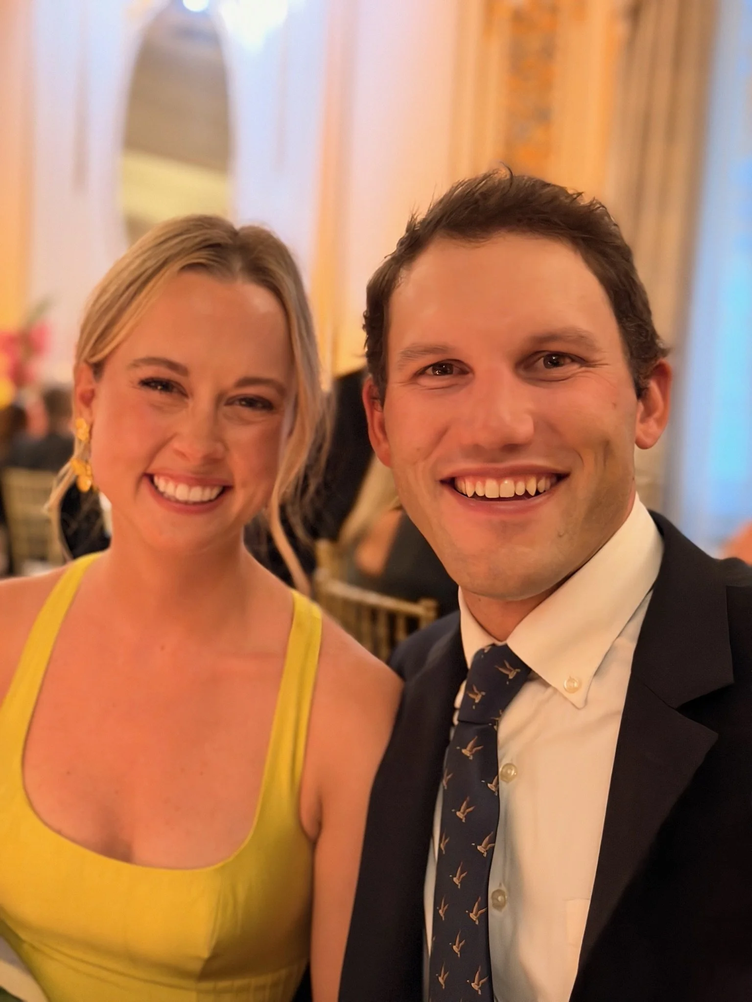 A smiling woman in a yellow dress and a smiling man in a black tuxedo with a patterned tie taking a selfie at a formal event.