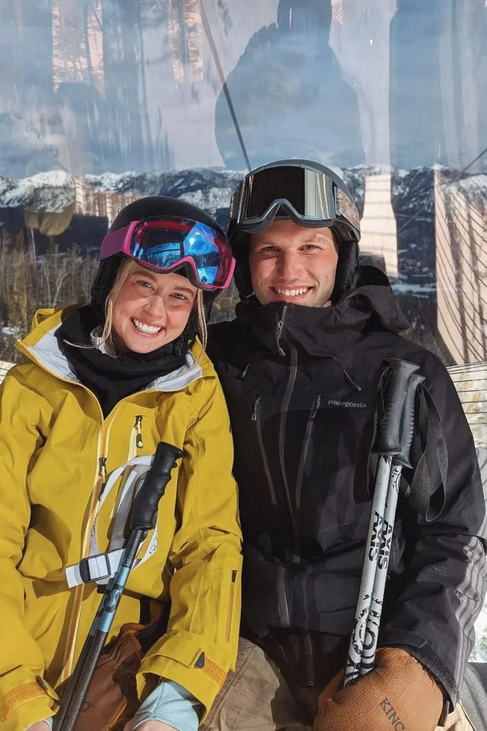Two smiling skiers, a woman in a yellow jacket and a man in a black jacket, wearing helmets and ski goggles, standing inside a ski lift cabin with snowy mountains visible in the background.