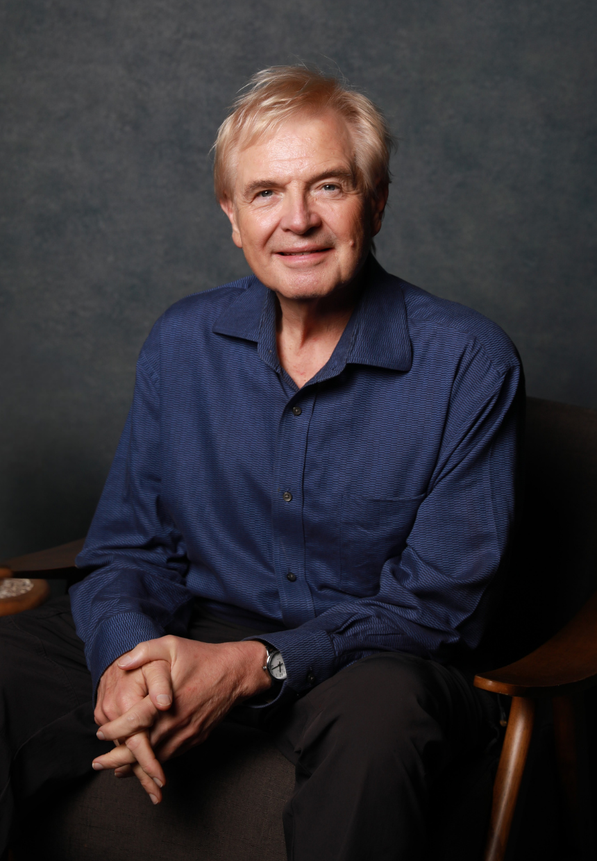 An older man with blond hair, wearing a blue long-sleeve button-up shirt, sitting on a dark-colored couch against a dark textured background.