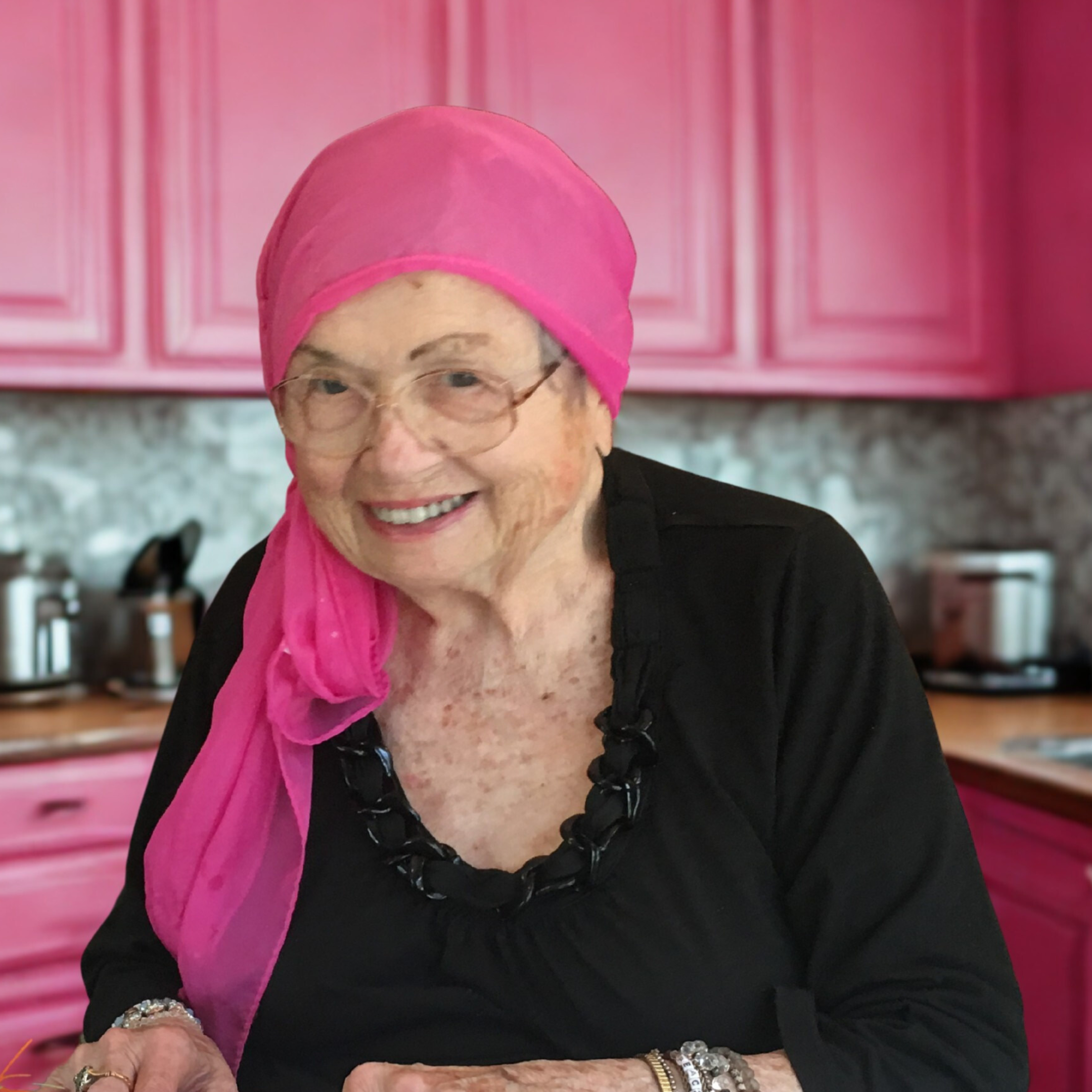 An elderly woman smiling in a kitchen with pink cabinets, wearing glasses, a pink headscarf, a black top, and jewelry.