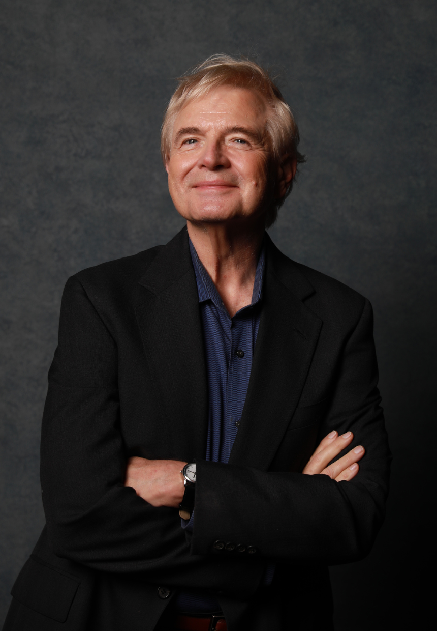 A confident elderly man with gray hair, wearing a black blazer and a blue dress shirt, standing with arms crossed against a dark textured background.