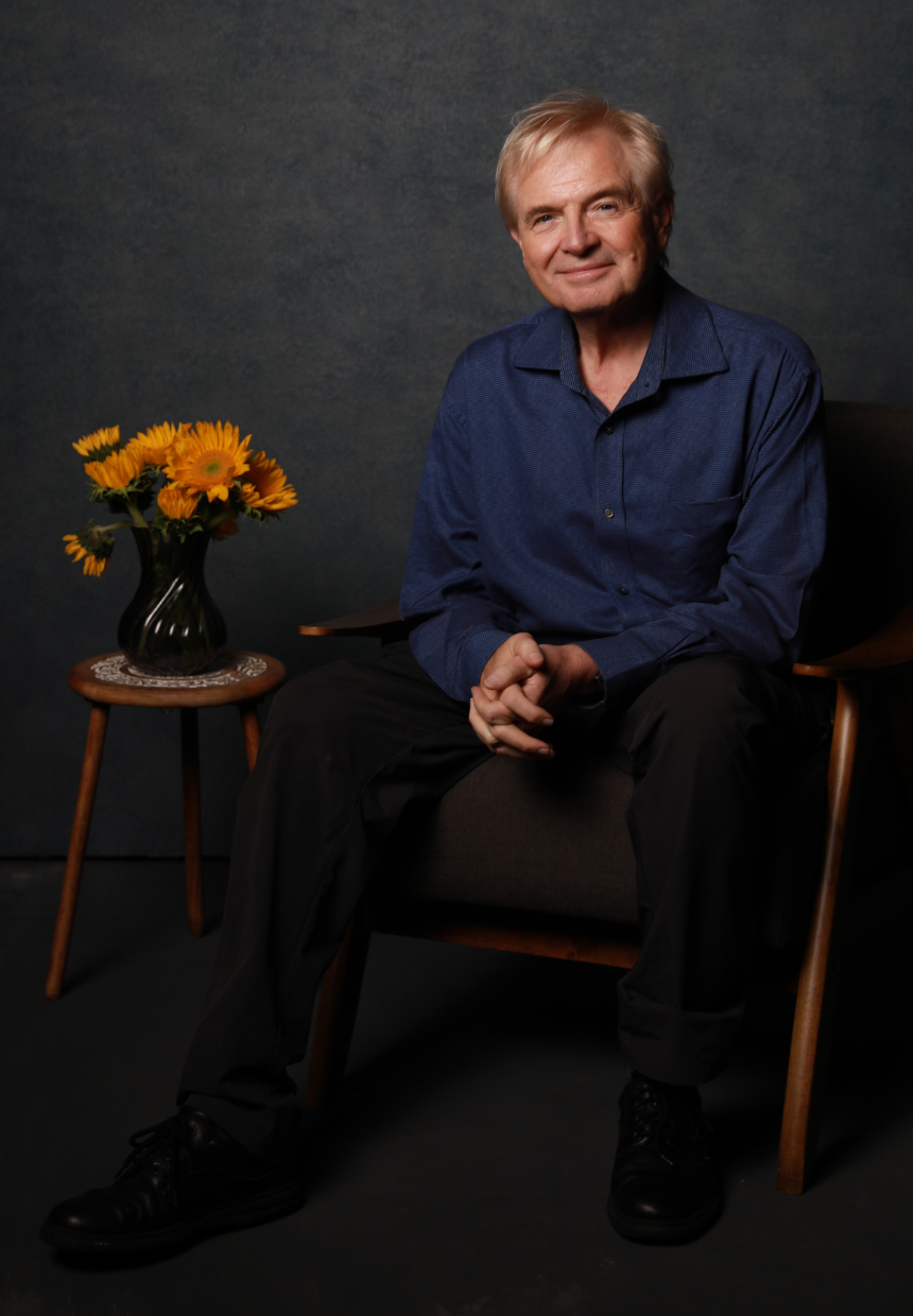 An elderly man with gray hair wearing a blue shirt sitting on a dark chair, with a small wooden table holding a black vase with yellow sunflowers next to him, against a dark background.