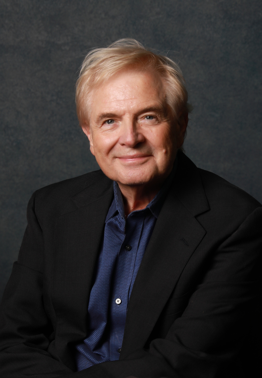 Portrait of an older man with blond hair, wearing a dark suit and a blue shirt, smiling against a dark background.