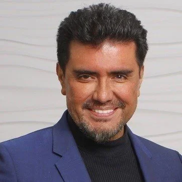 A man with dark hair and a beard, smiling, wearing a Navy blue blazer and black shirt, standing in front of a white wooden background.