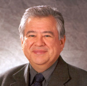 A professional headshot of a middle-aged man with gray hair, wearing a dark suit and tie, smiling against a neutral background.