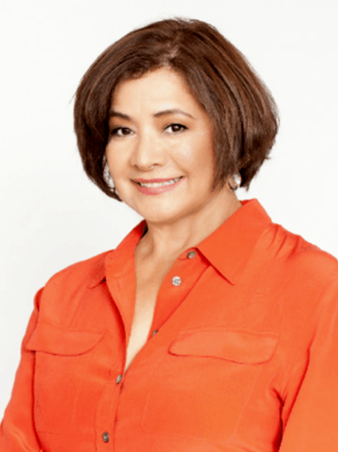 A woman with short brown hair wearing an orange button-up shirt, smiling at the camera against a white background.