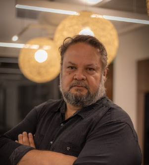 A man with a beard and curly hair standing indoors with arms crossed, looking at the camera. In the background, there are hanging light fixtures and decorative lighting.
