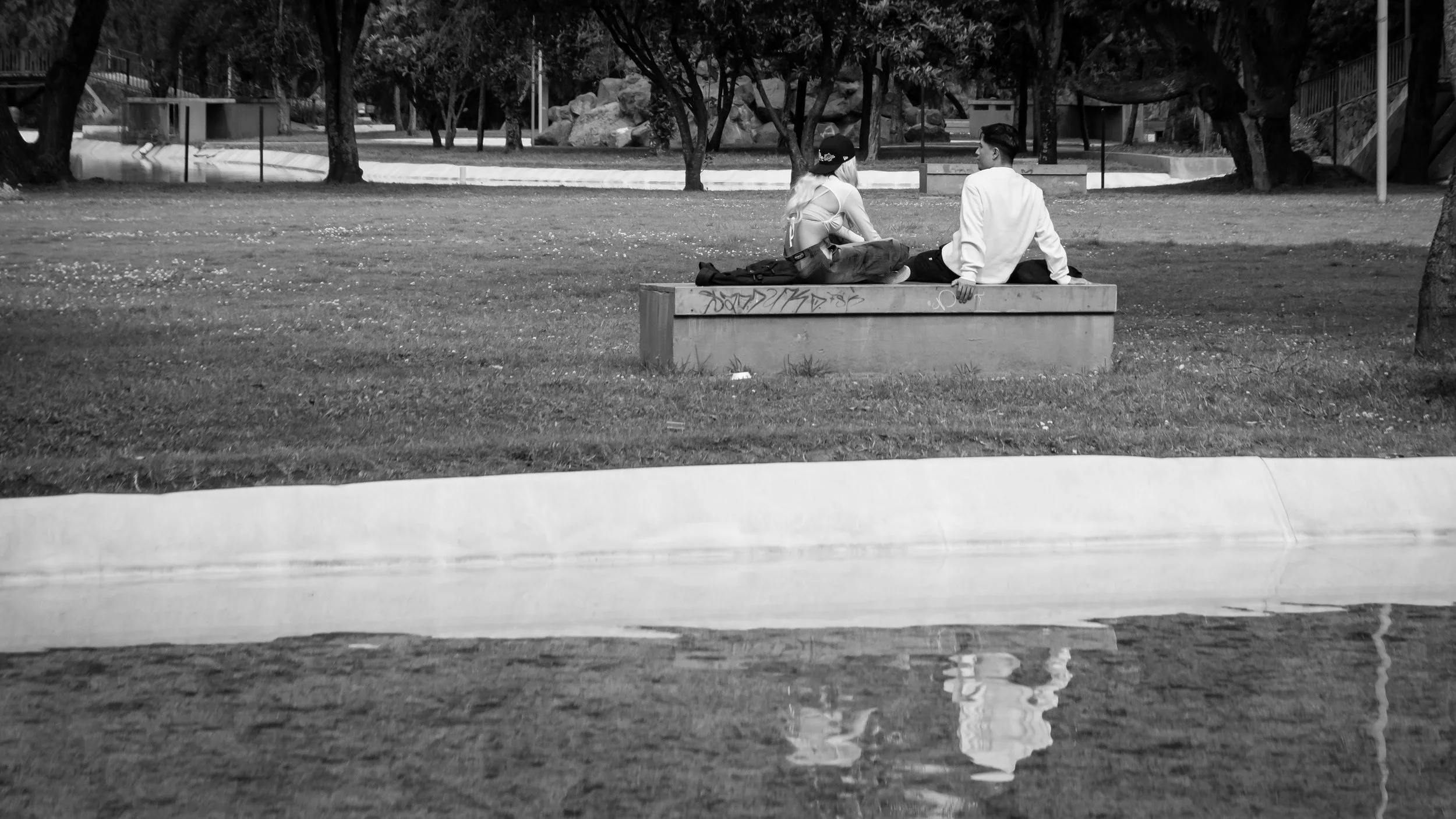 Two young people sitting on a park bench, engaged in conversation, with trees and rocks in the background, reflected in a water puddle on the ground.