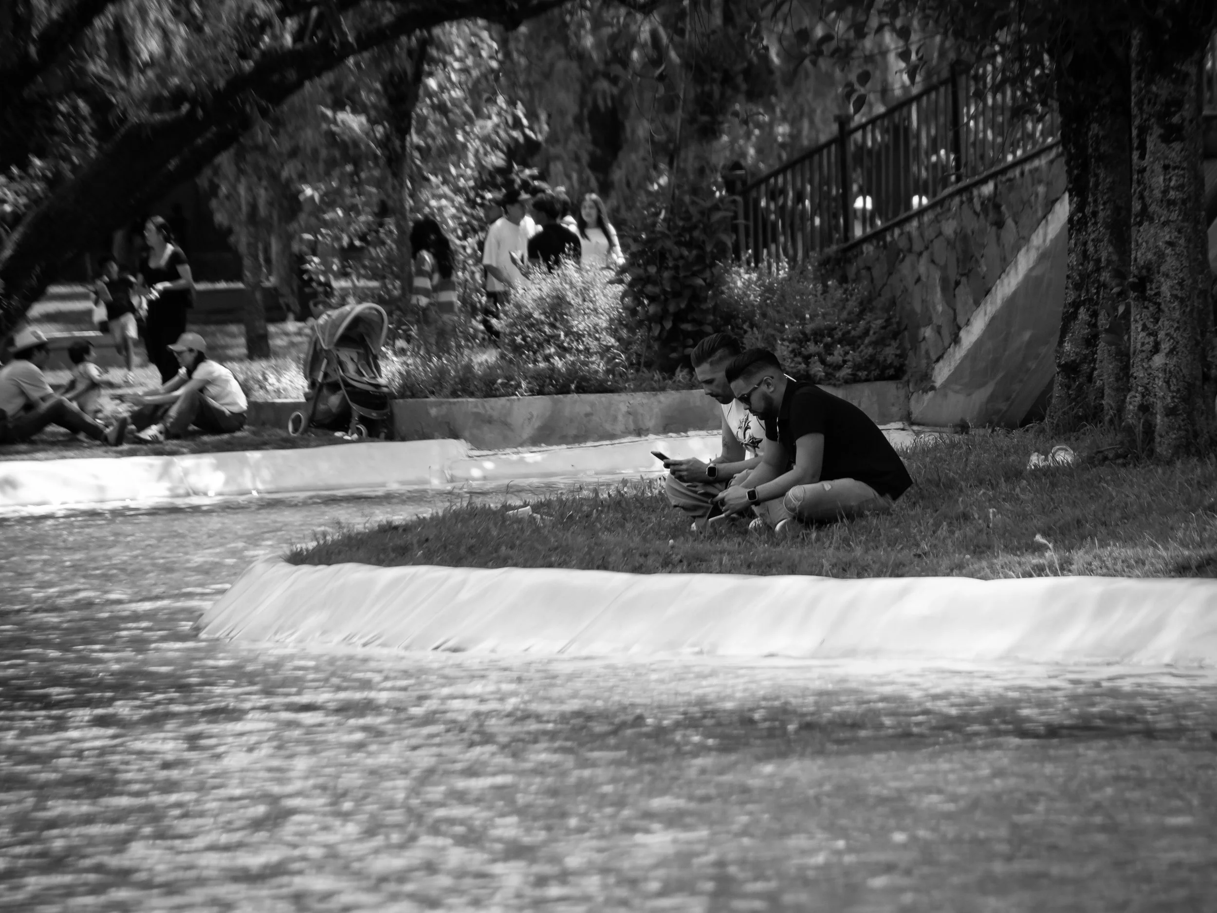 Black and white photo of people sitting and chatting outdoors beneath trees, with some standing and walking in the background, and a metal fence on the right.