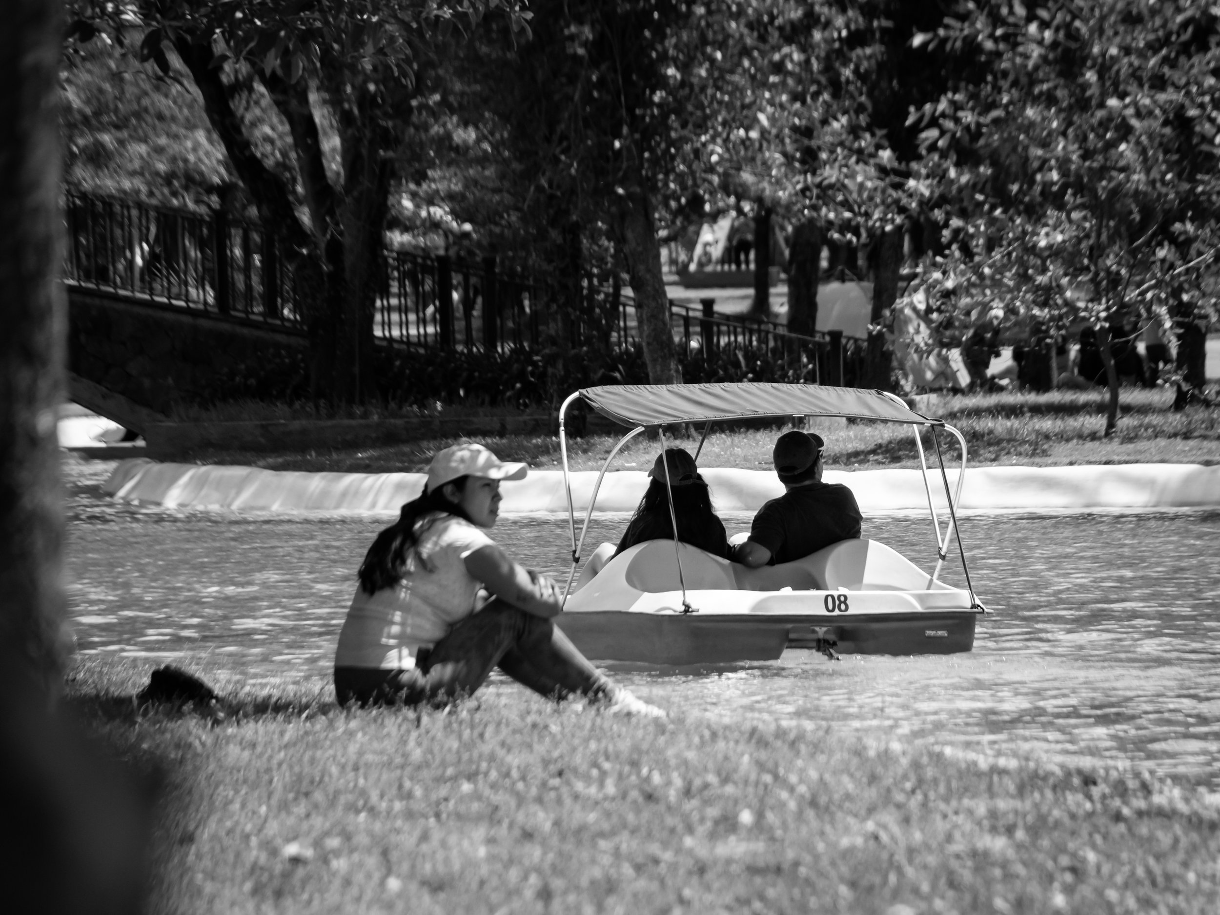 Three people sitting in a small boat on a river, with one person in the foreground sitting on the grassy riverbank, and a row of trees and a fence in the background. It is a black and white photograph.