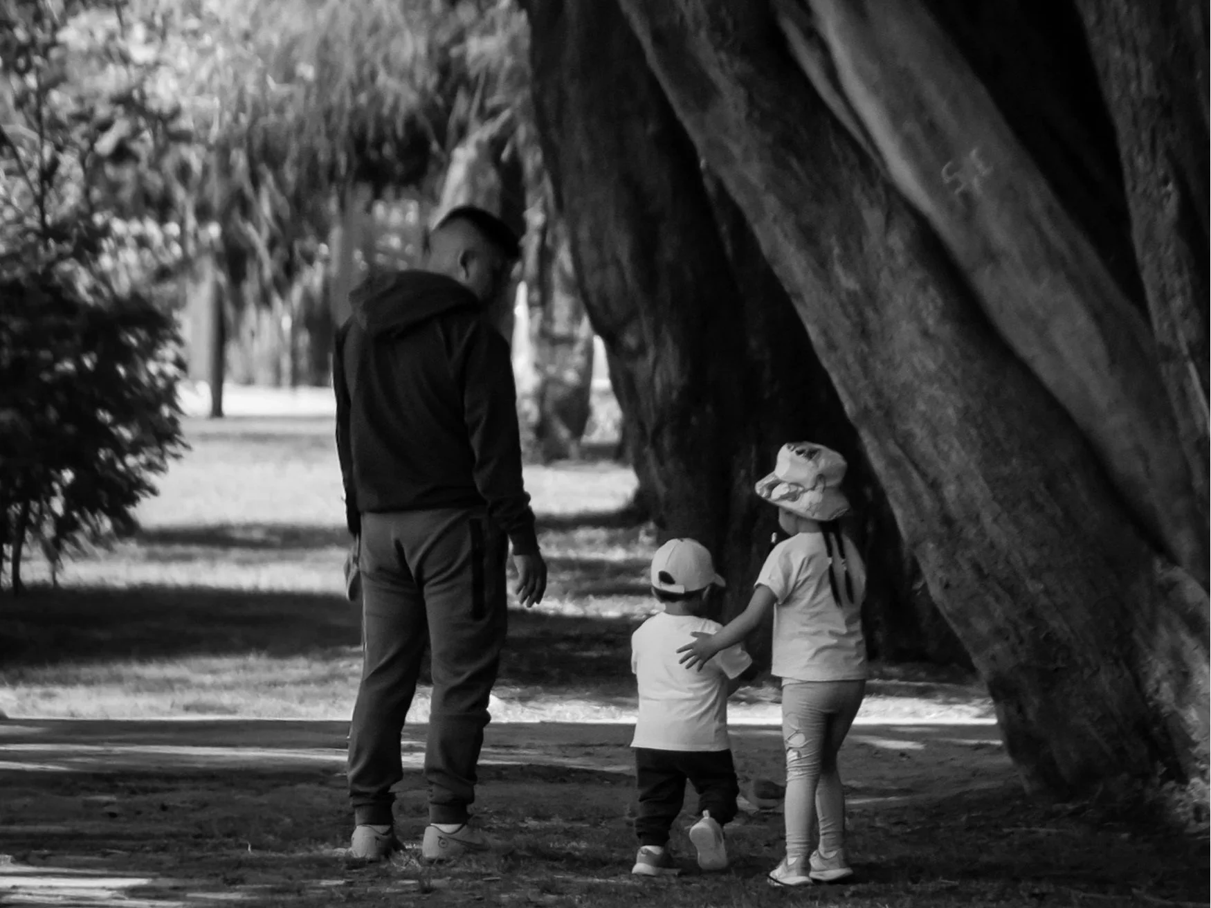 A man and three children standing and sitting under a large tree in a park or yard, with some of the children wearing hats and casual clothing.