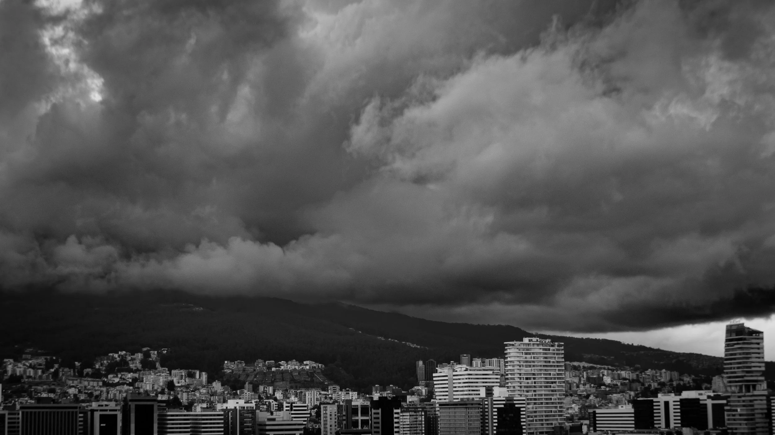Black and white photo of a city skyline with tall buildings and dark, ominous clouds overhead, indicating an approaching storm.