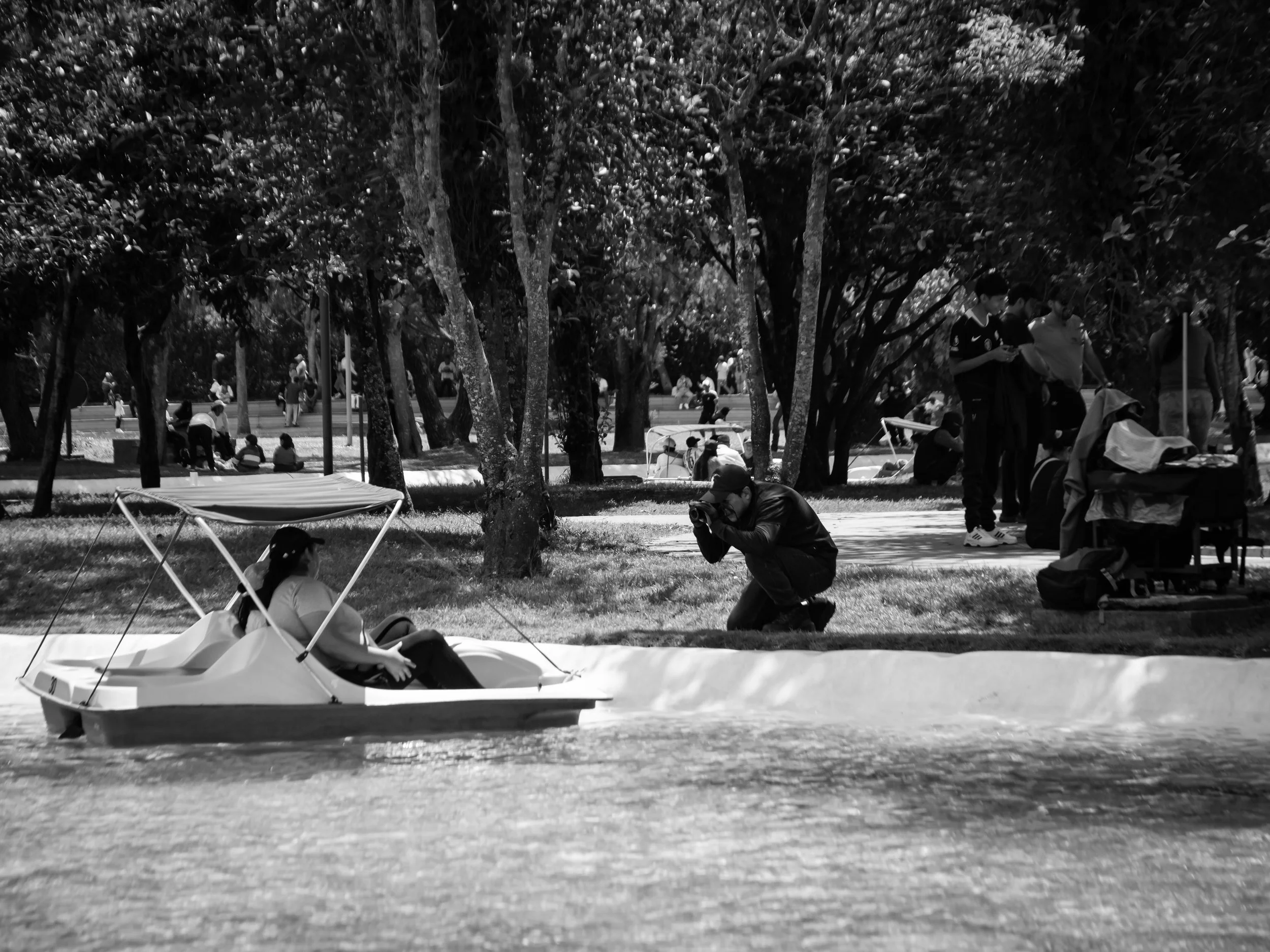 Black and white photo of a park with many trees and people sitting on the grass or walking. A small boat is in the water in the foreground, with a person leaning on the boat.