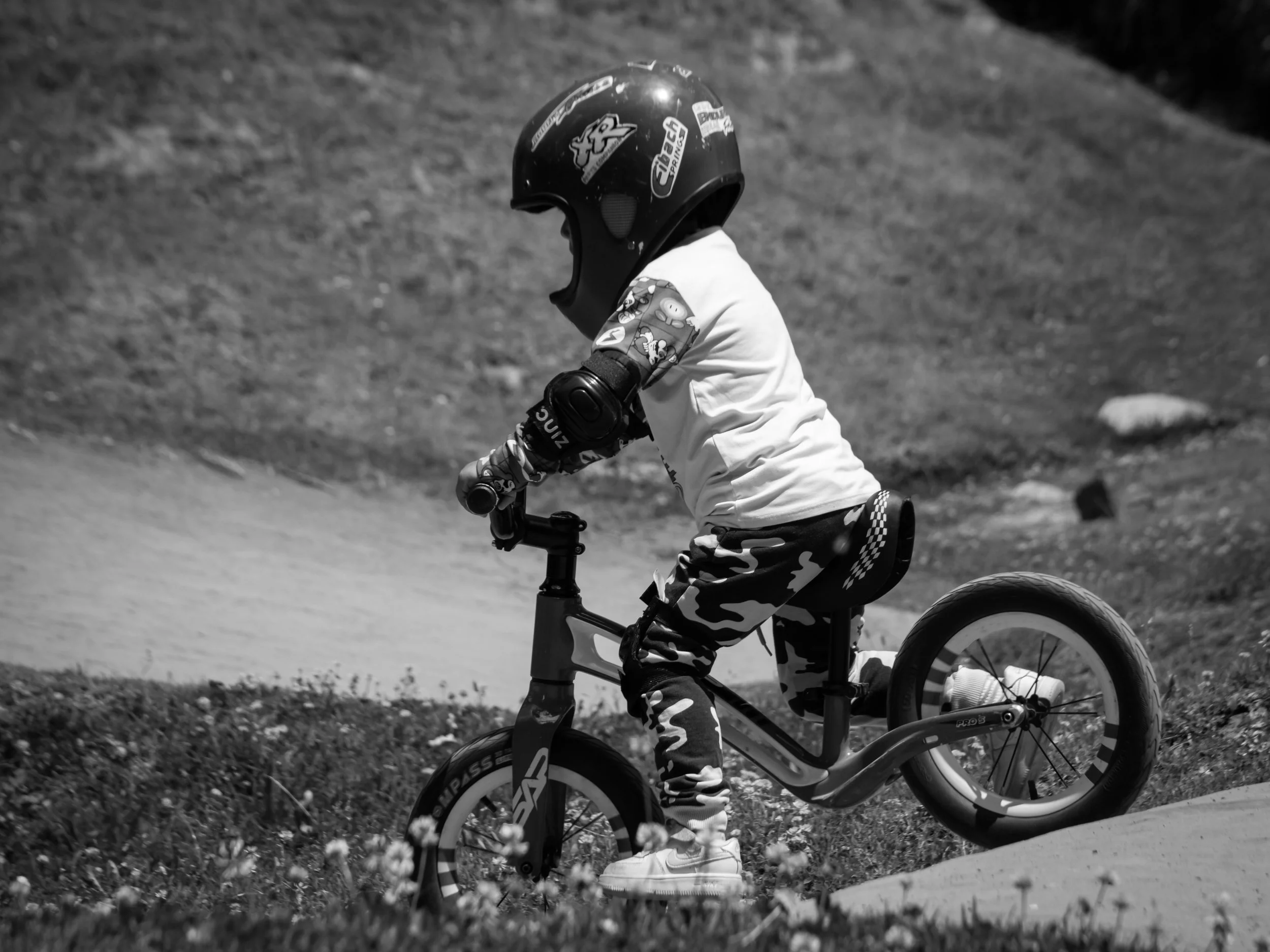 A young child riding a bicycle outdoors, wearing a helmet and camouflage-patterned pants. The scene is in black and white.