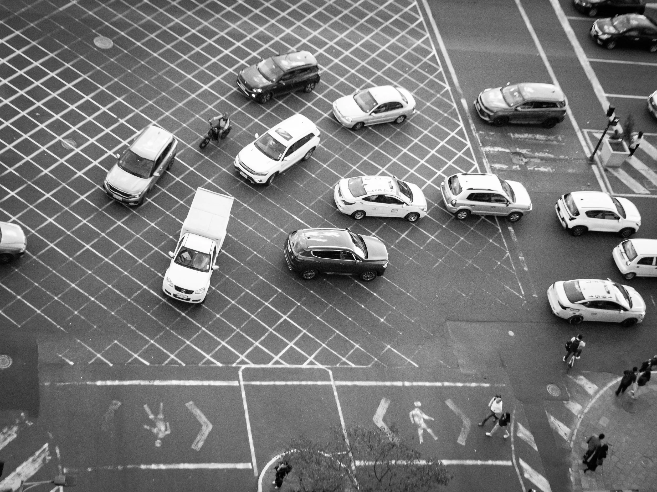Black-and-white aerial view of a parking lot with designated parking spaces, some of which are occupied by cars and a few by bicycles. A person walking nearby.