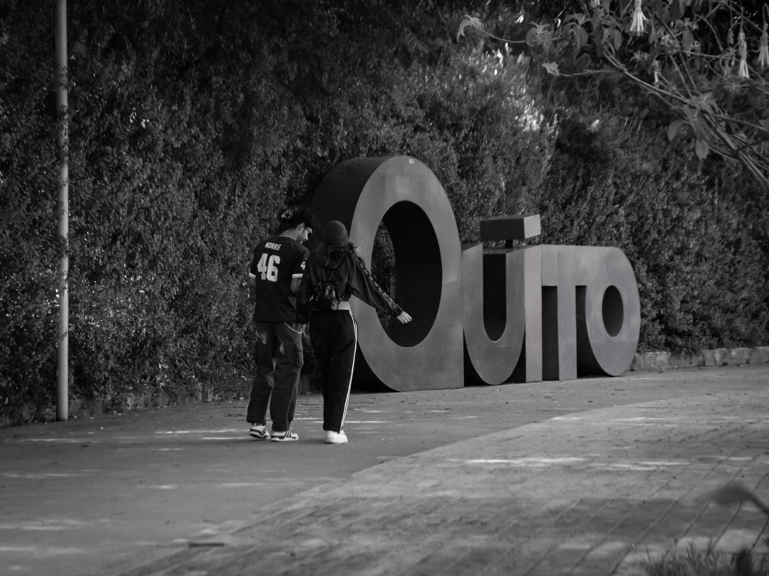 Two people walking together in an outdoor area at night, with large block letters spelling 'LOVE' in the background and trees behind them.