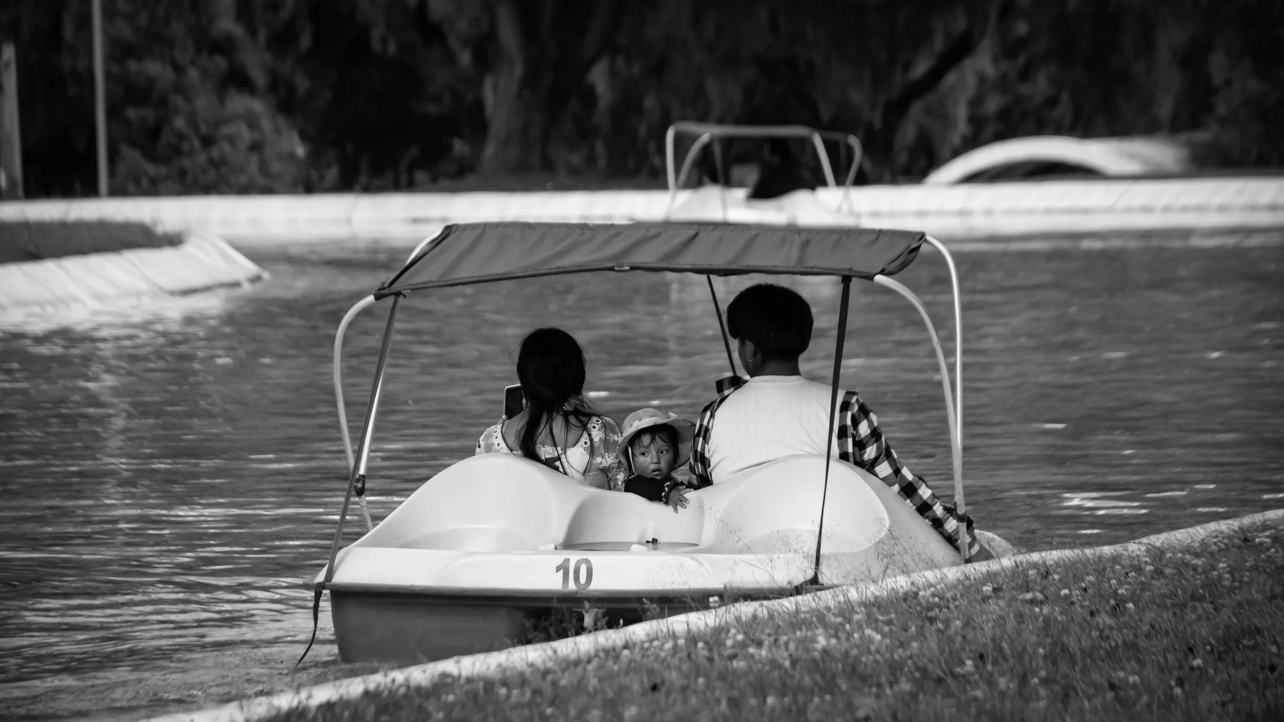 A family of three on a pedal boat on a river, with two children and one adult, all wearing casual clothes. The boat has a canopy, and the family appears to be enjoying a boat ride.