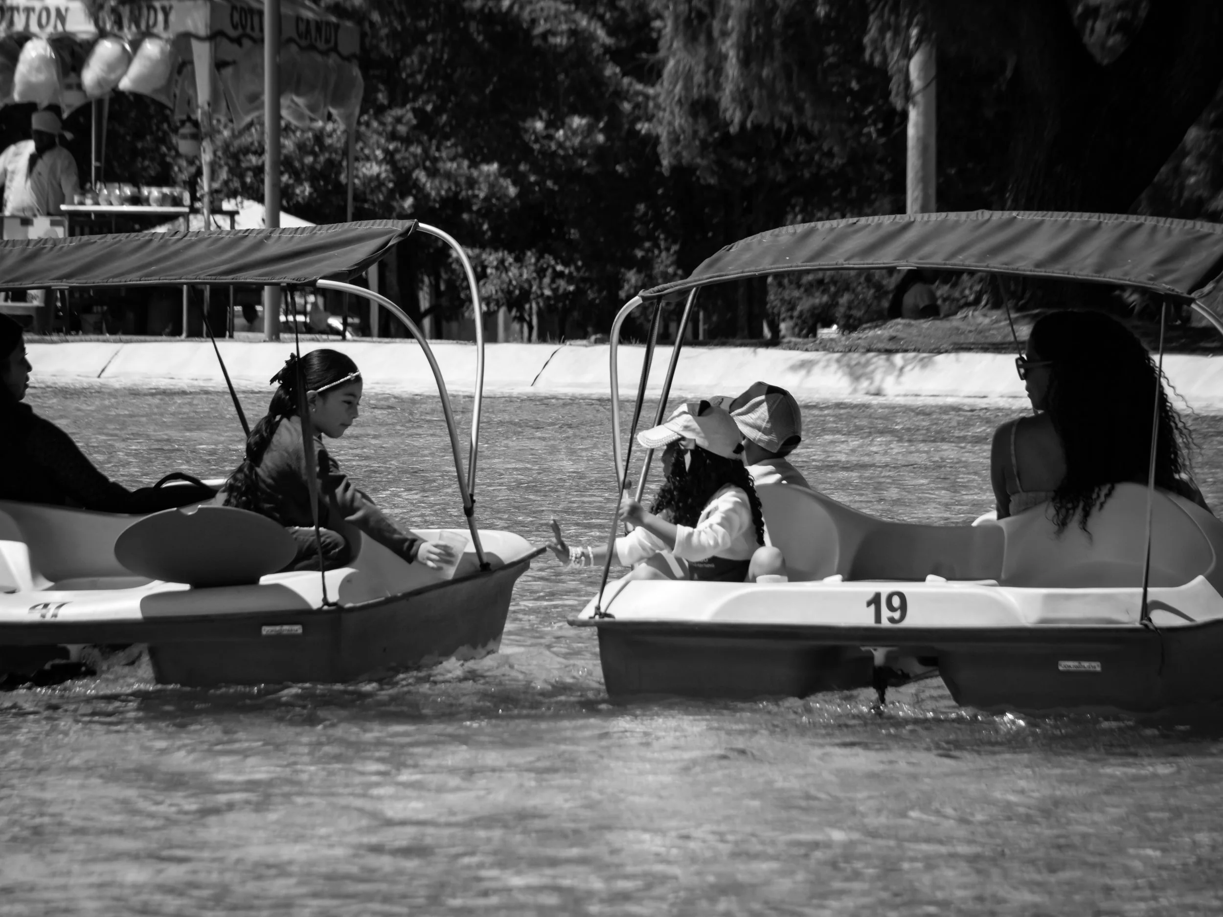 People on pedal boats in a lake or pond, some wearing hats, with trees in the background.