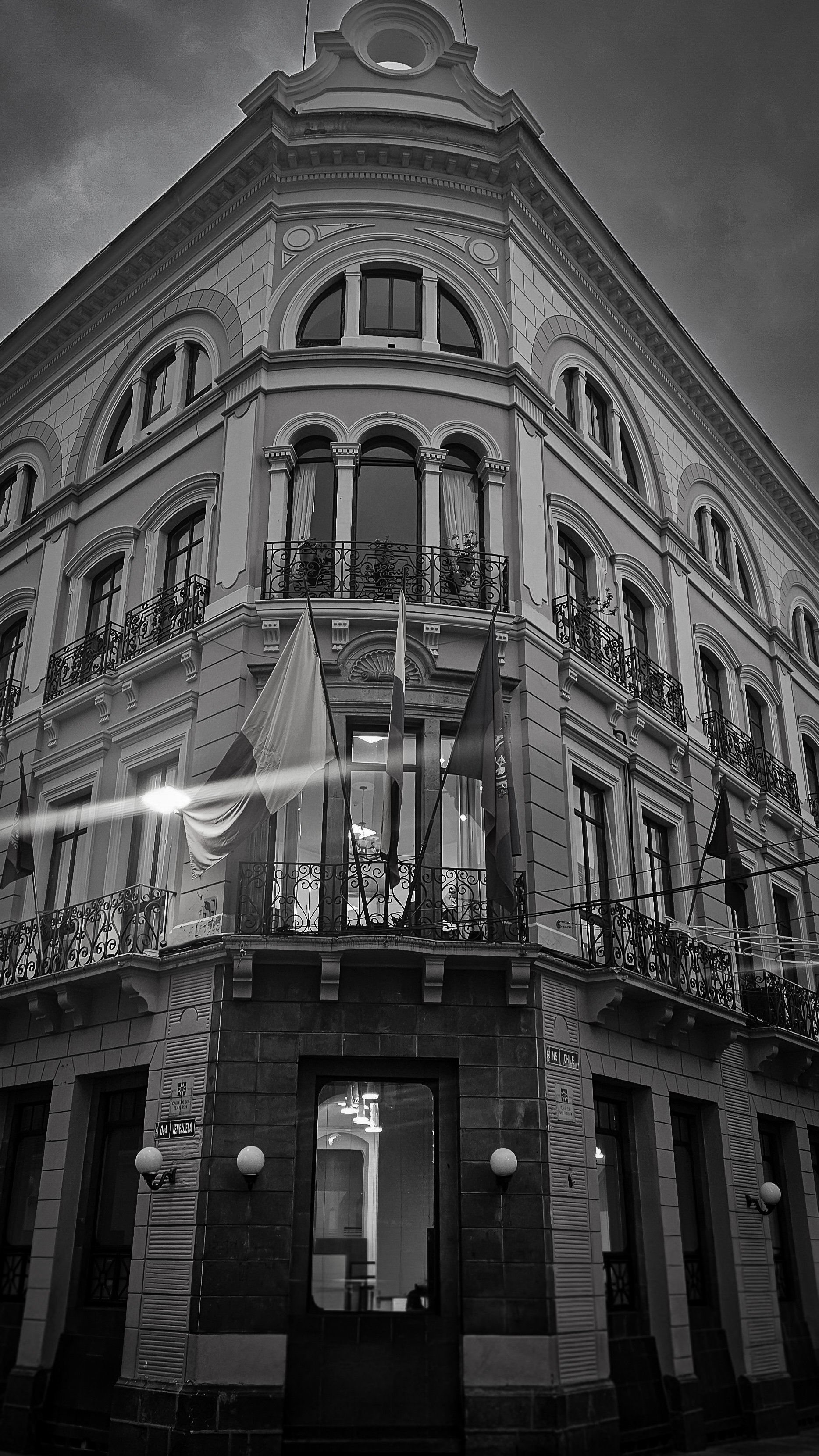 Black and white photo of a grand, ornate building on a street corner with flags hanging from the windows.