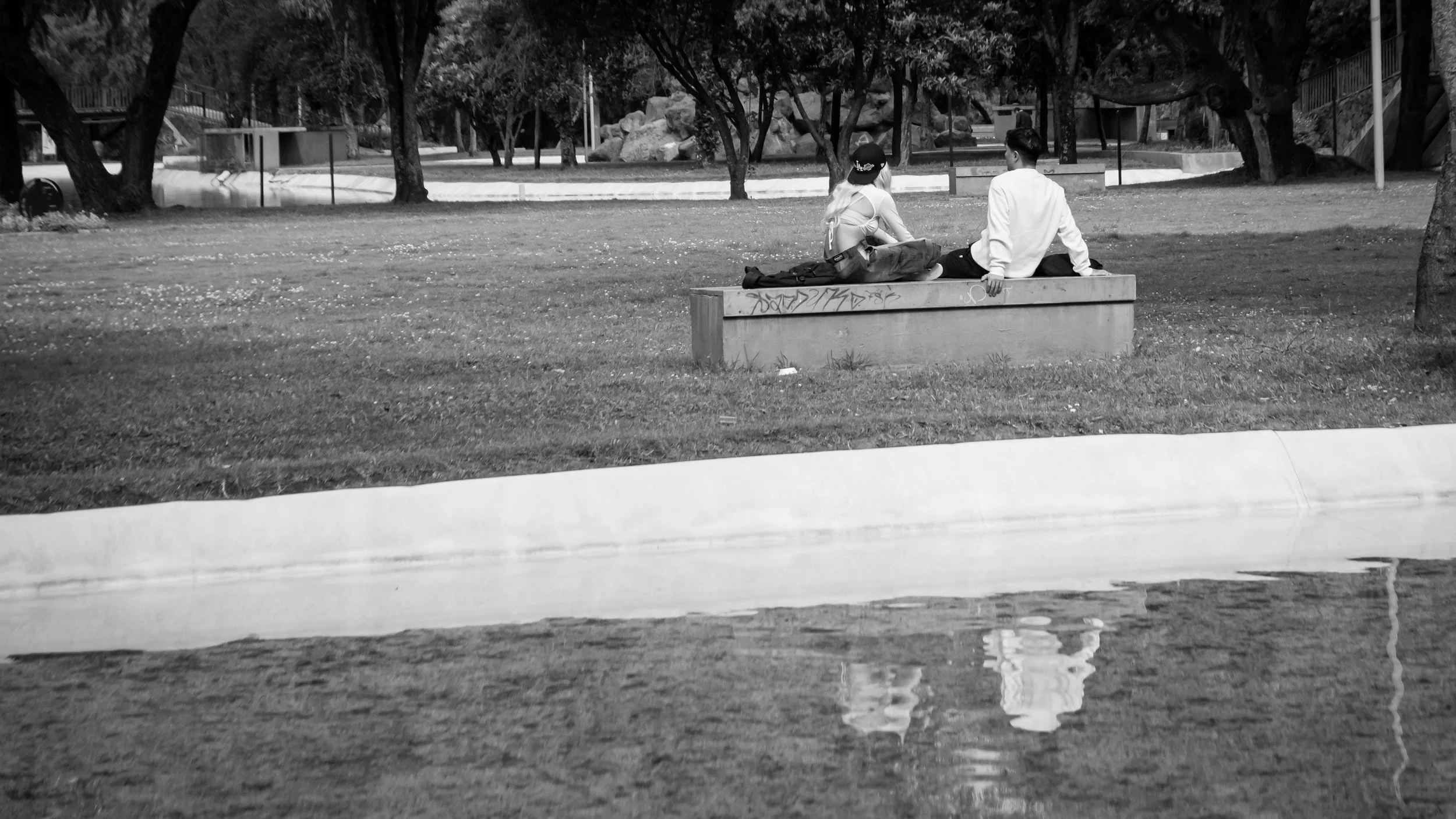 Two people sitting on a park bench near a body of water, surrounded by trees, in black and white.