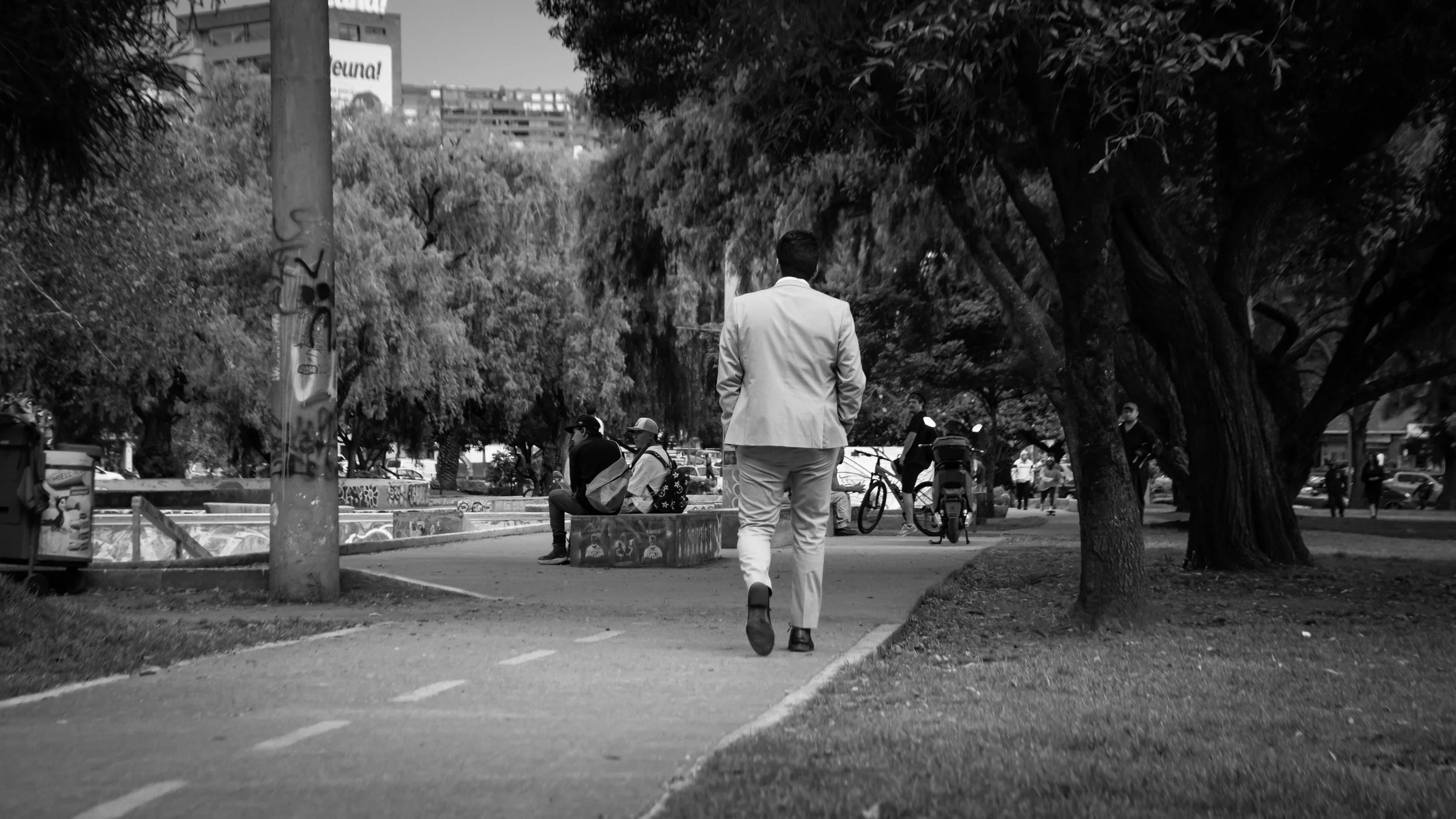 A man in a light-colored suit walking in a park with trees, other people sitting and standing, some with bicycles, and city buildings in the background.