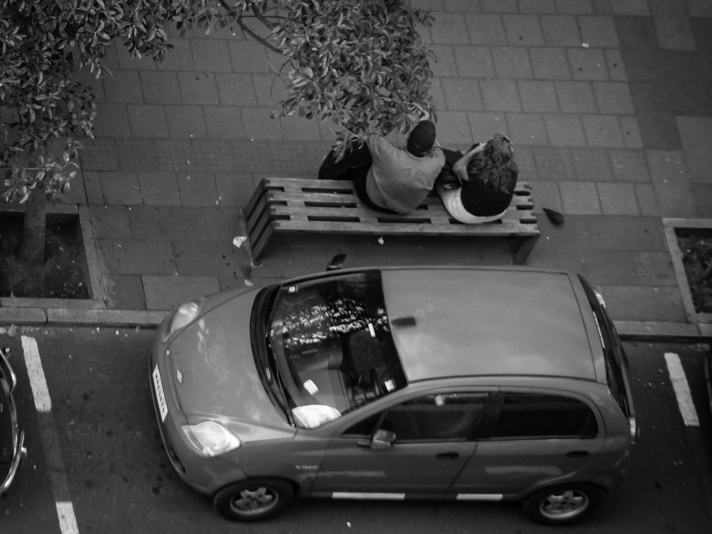An overhead view of a sidewalk with a wooden bench, two people sitting on the bench, a small tree, and parked cars along the curb.