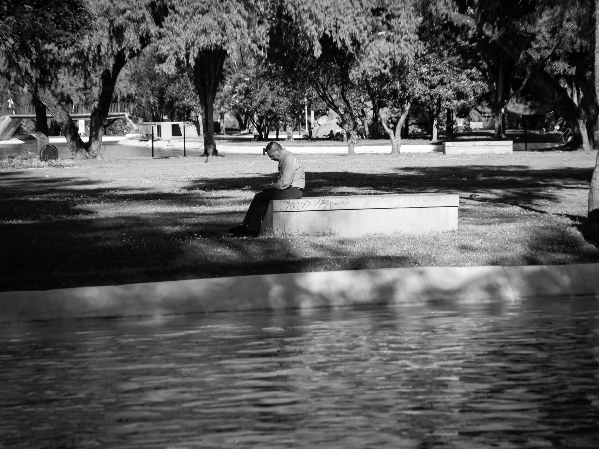 A man sitting on a concrete bench in a park, surrounded by trees and shaded areas, black and white photograph.