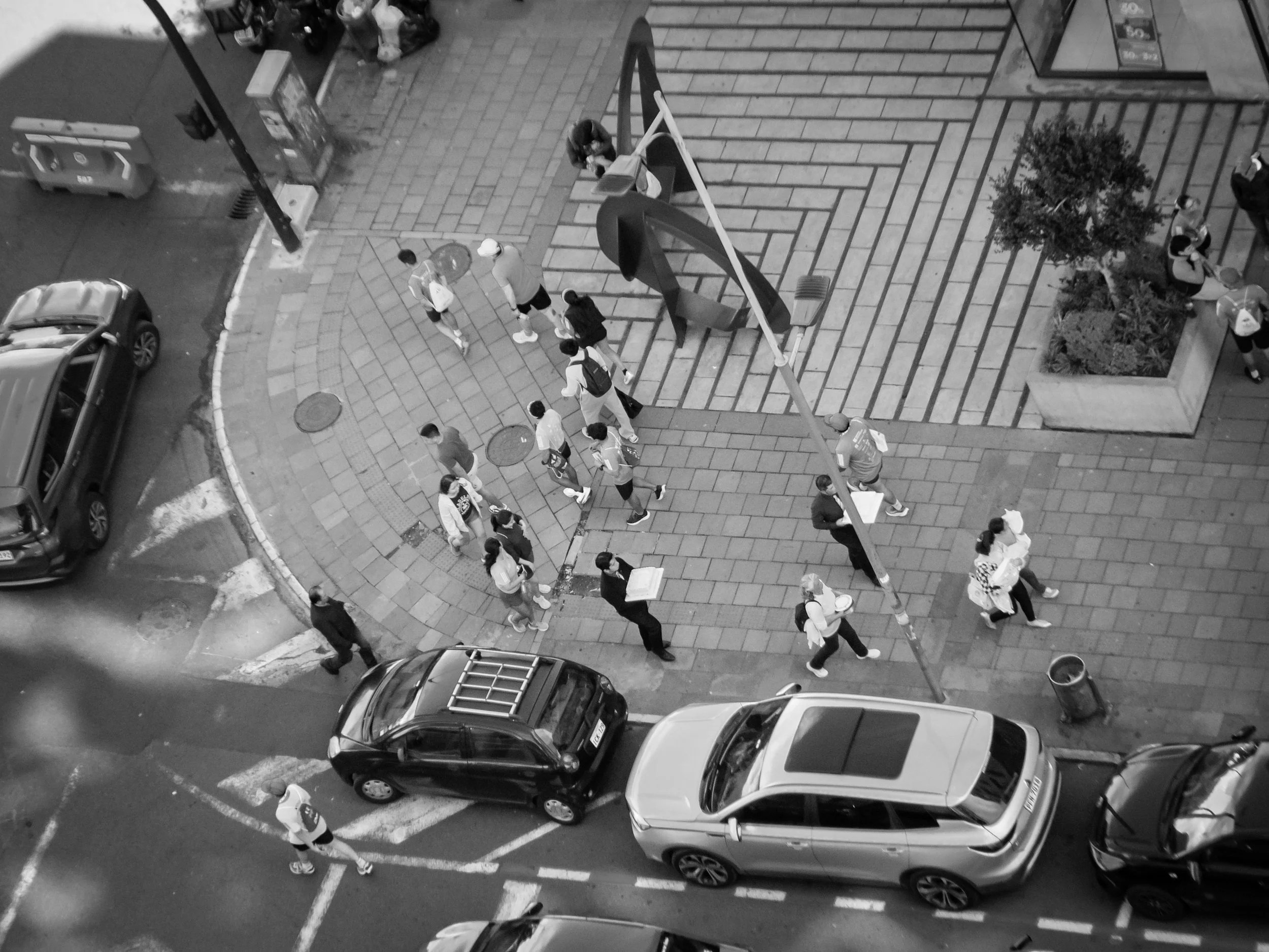 A black and white aerial view of a city street corner showing parked cars along the curb, people walking on the sidewalk, and a street basketball hoop.