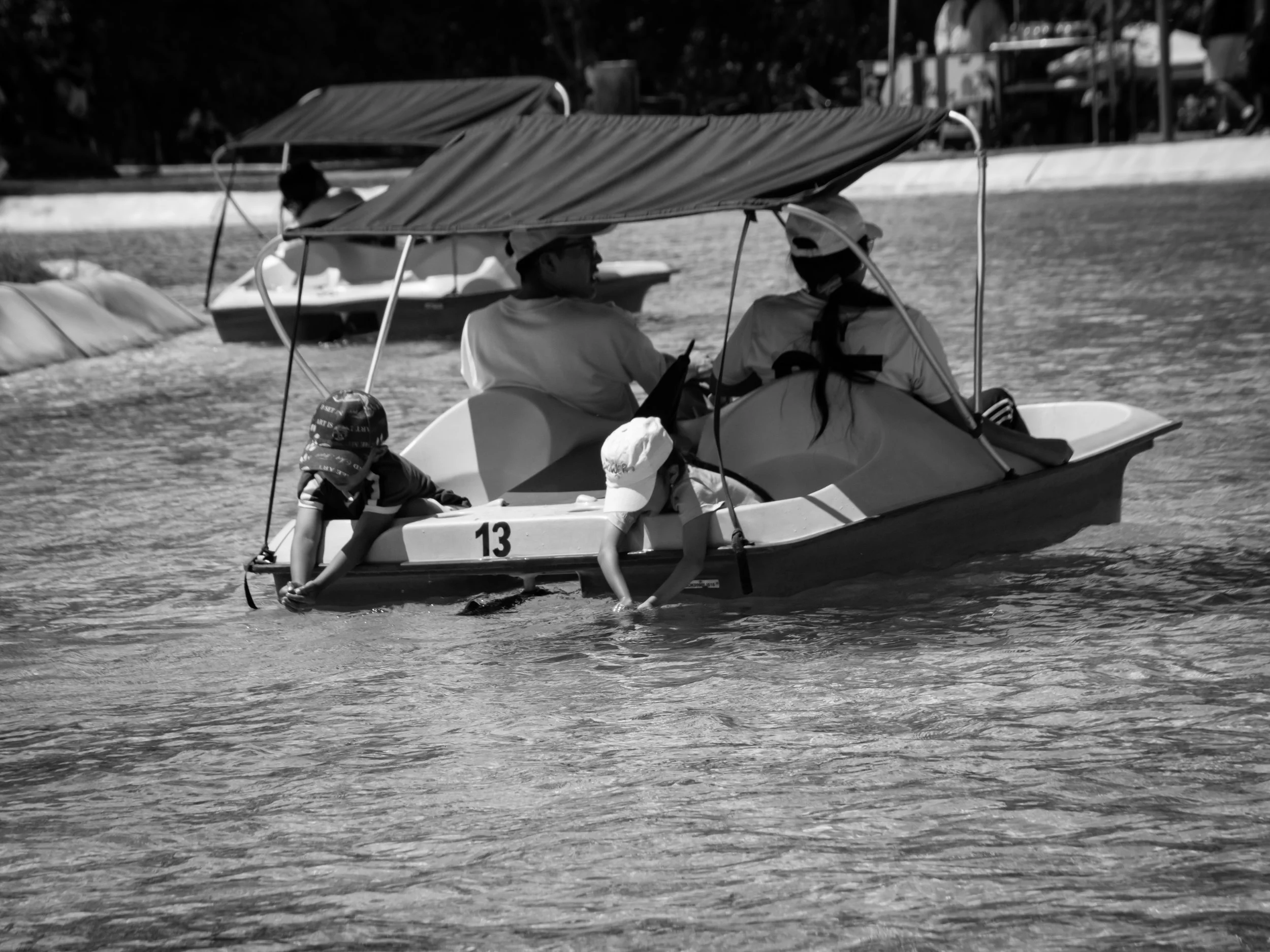 People on pedal boats with canopy shades on a body of water, children sitting at the front of one boat, and additional boats and shoreline in the background.