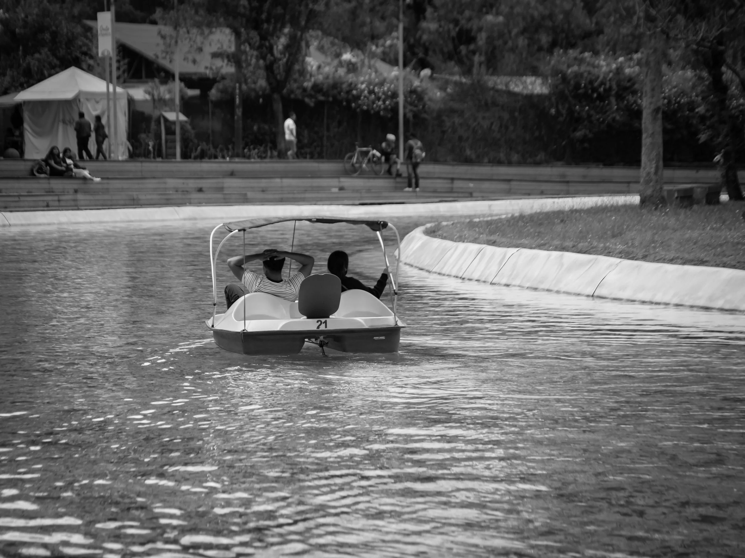 A black-and-white photo of two people sitting in a pedal boat on a body of water with a curved shoreline, with several people walking or biking on a path and trees in the background.