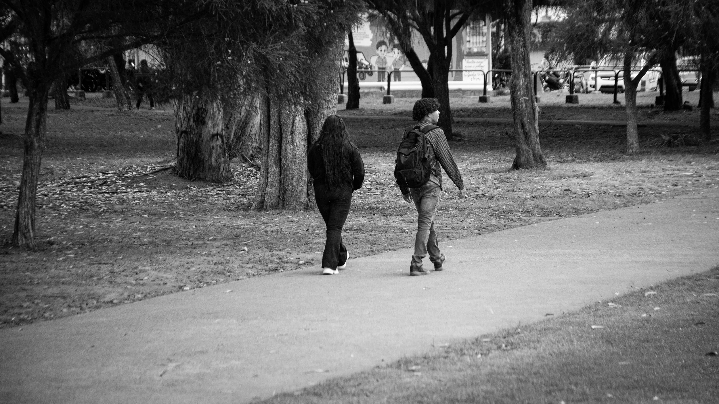 A black and white photo of a young man and woman walking on a sidewalk near trees in a park-like area. The woman has long hair and is wearing dark clothing, while the man has curly hair, a backpack, and jeans. In the background, there are more trees,