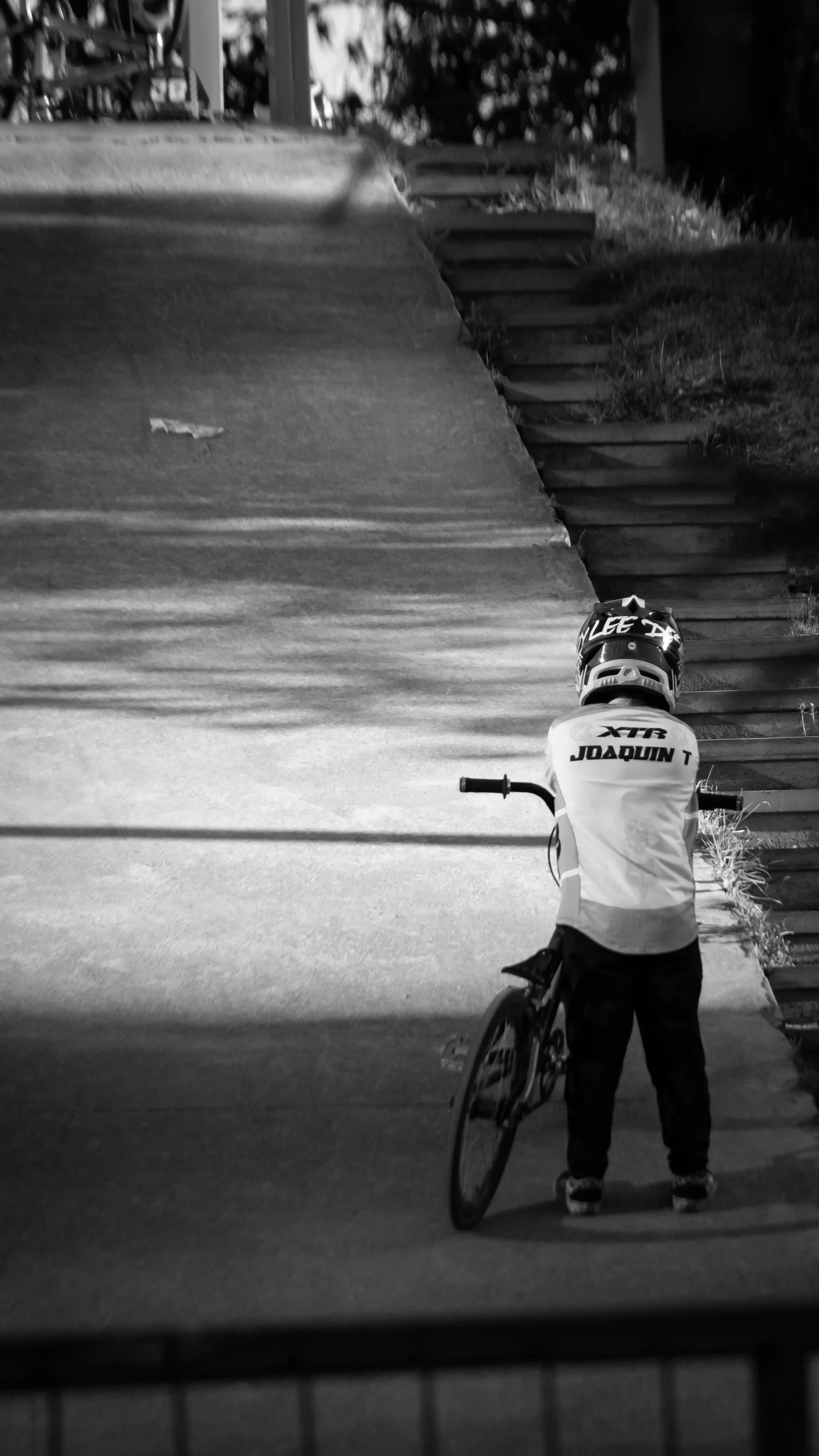 A person wearing a helmet and a jersey with the name 'Joaquin' on the back, standing with a bicycle in front of a staircase outdoors, captured in black and white.